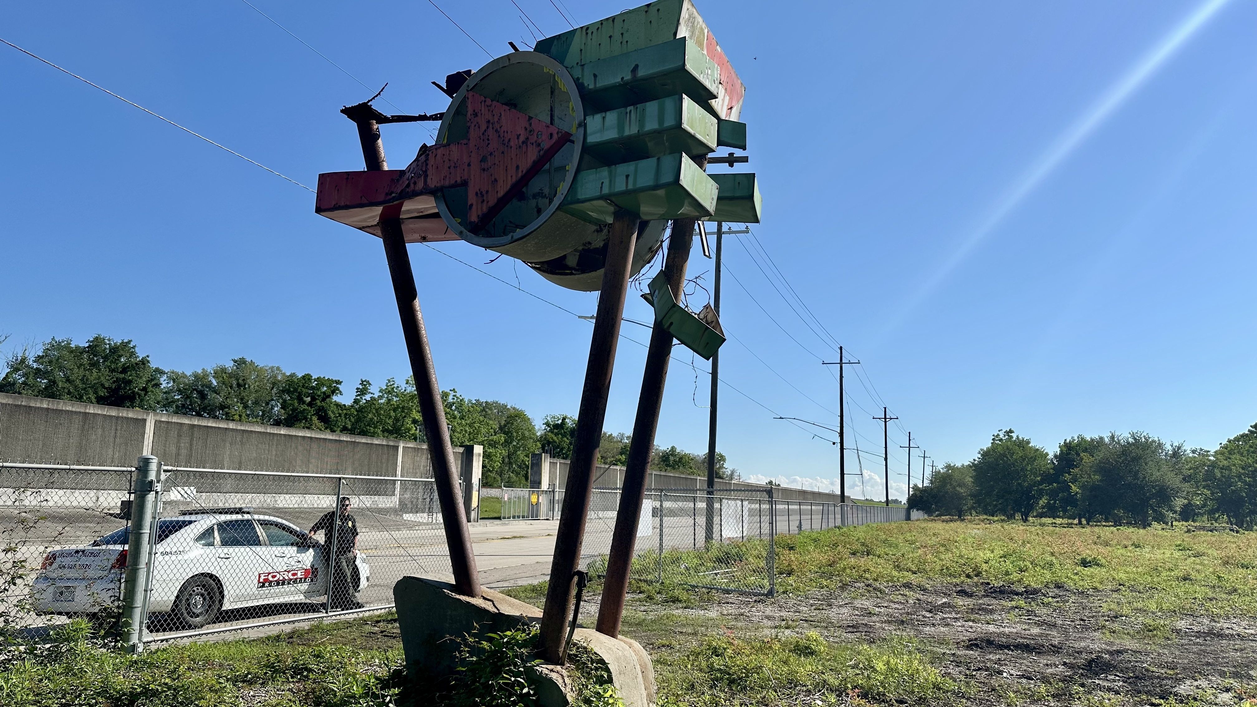 Rusty, broken green and red arrow sign on metal poles beside a fenced lot with grass, trees, and a road under a clear blue sky during daytime, with a security car and guard nearby.