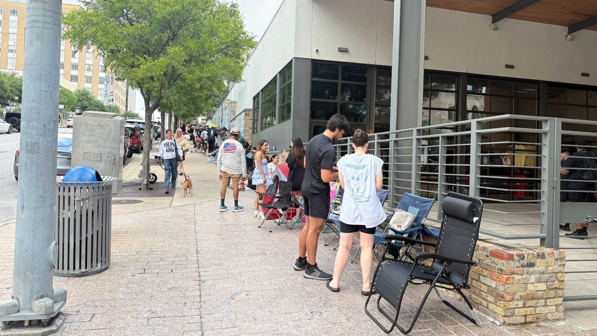 Urban sidewalk scene with people lined up along a modern building, folding chairs set outside, a large tree nearby, and a red pedestrian signal in the foreground.