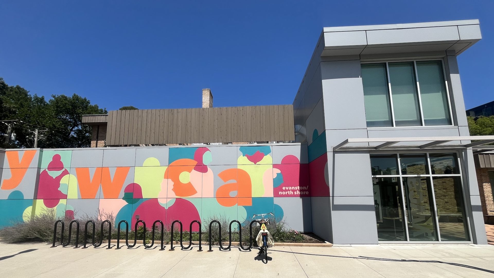 Colorful mural with abstract shapes and oversized letters on a wall in front of a modern gray building with large glass windows; a bicycle rests by a black looping bike rack under a clear blue sky.