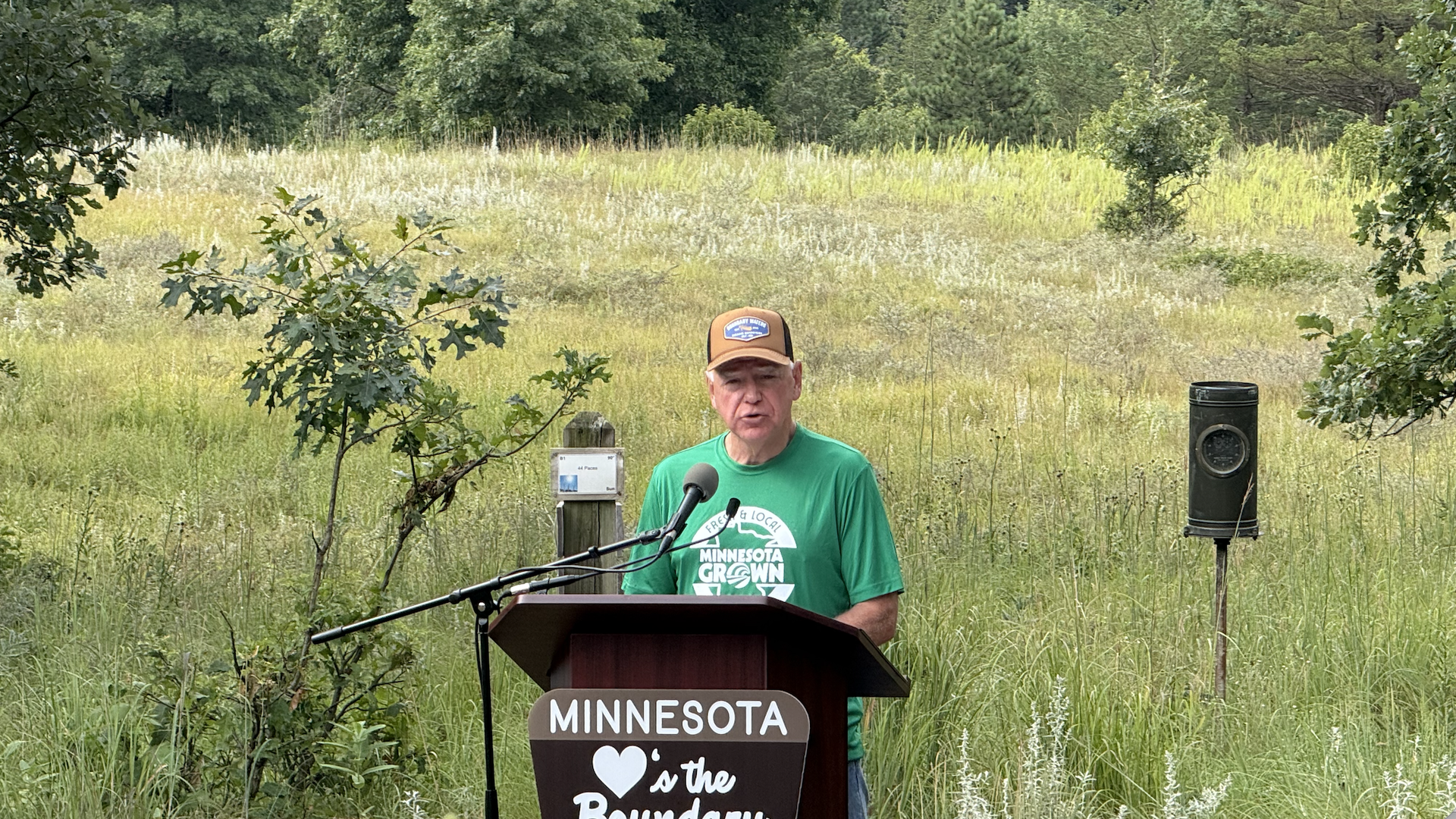 An older man in a green shirt and brown cap stands at a podium outdoors, speaking into a microphone in a grassy field with trees in the background and a Minnesota sign on the podium.