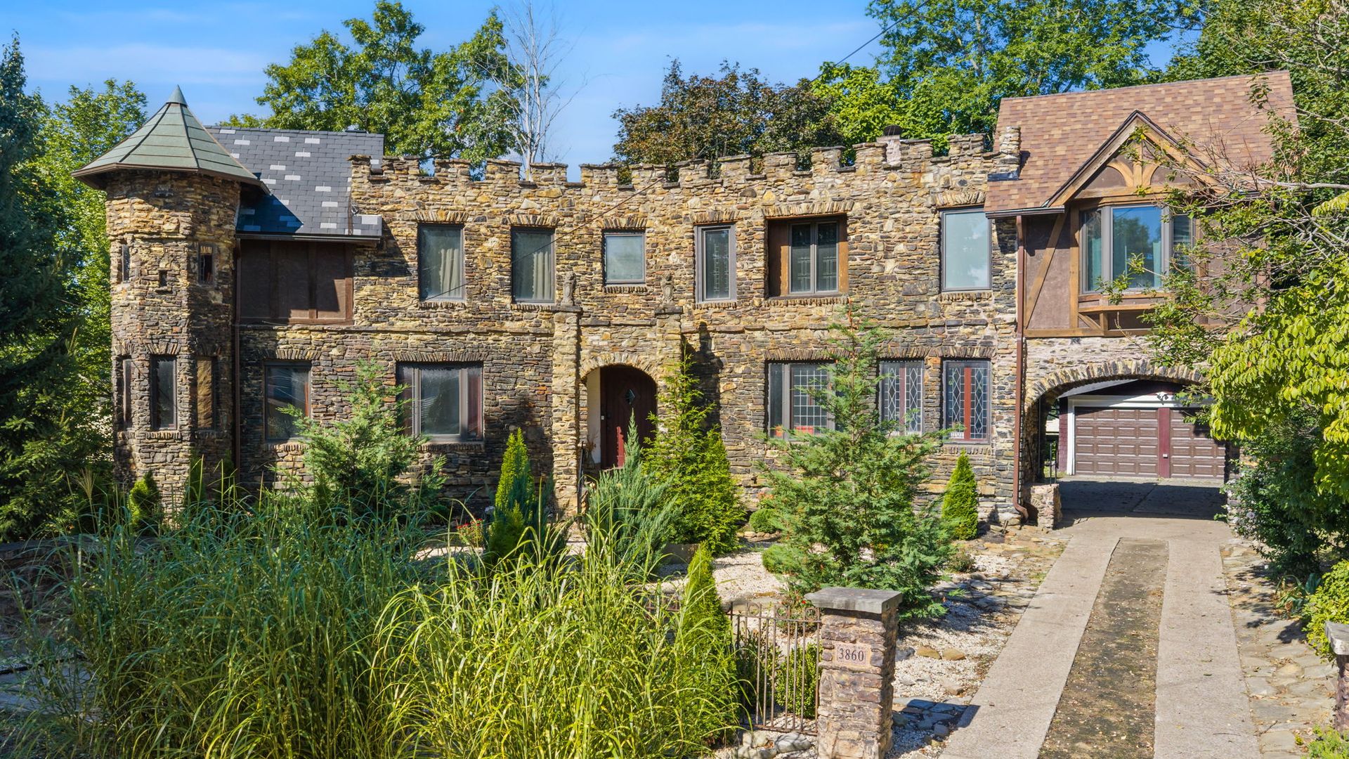 Stone mansion with a crenellated roofline and a turret on the left, multiple tall windows, a brown-roofed section on the right, and lush landscaping under a bright blue sky.