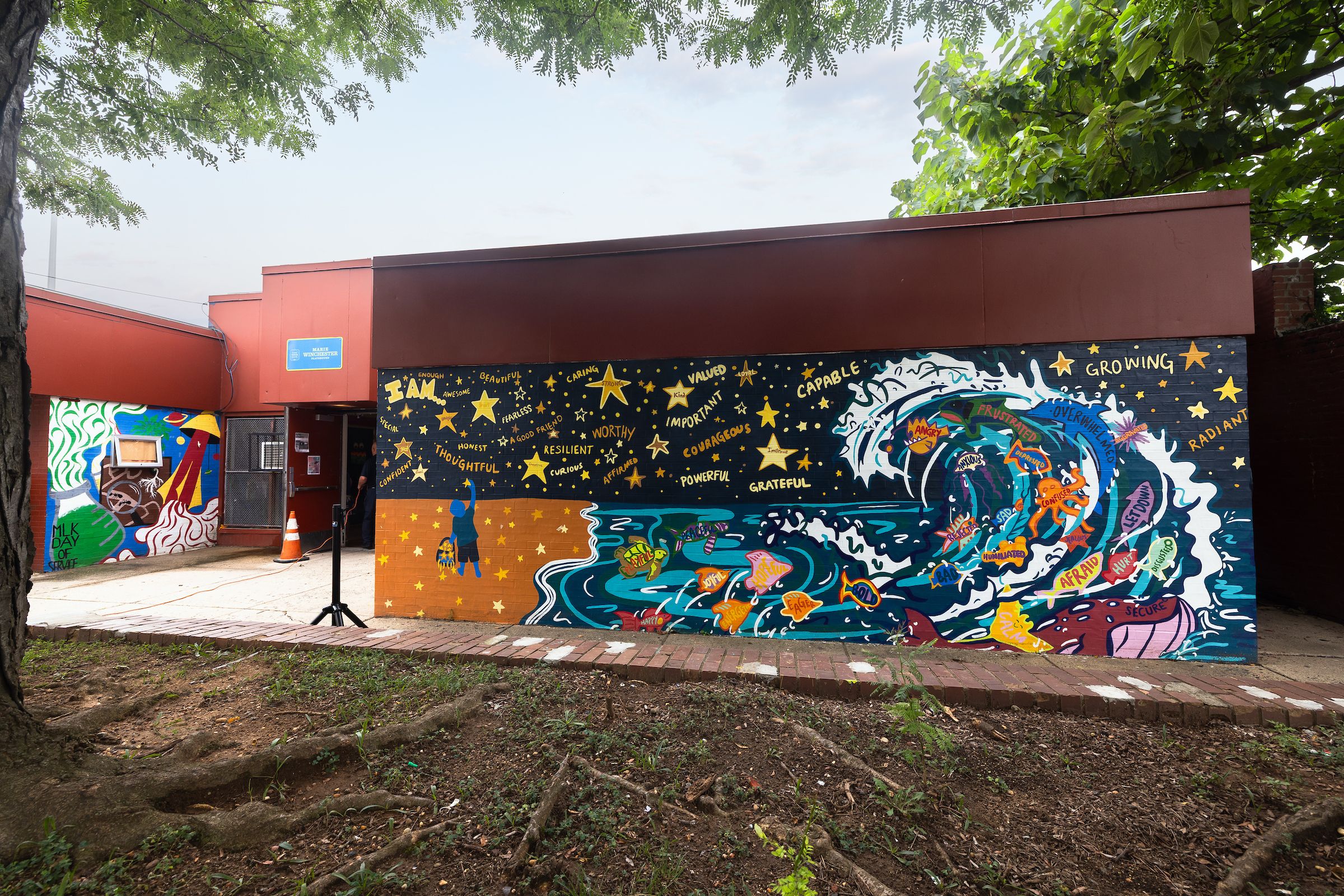 A beach mural on the side of a low-slung building featuring a wave with sea animals, including octopuses and fish, with inspirational words written on them, along with words written in the sky and stars. 