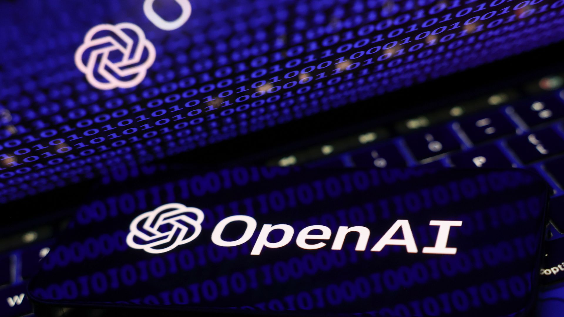 Image of binary code displayed on a laptop screen and OpenAI logo displayed on a phone screen.