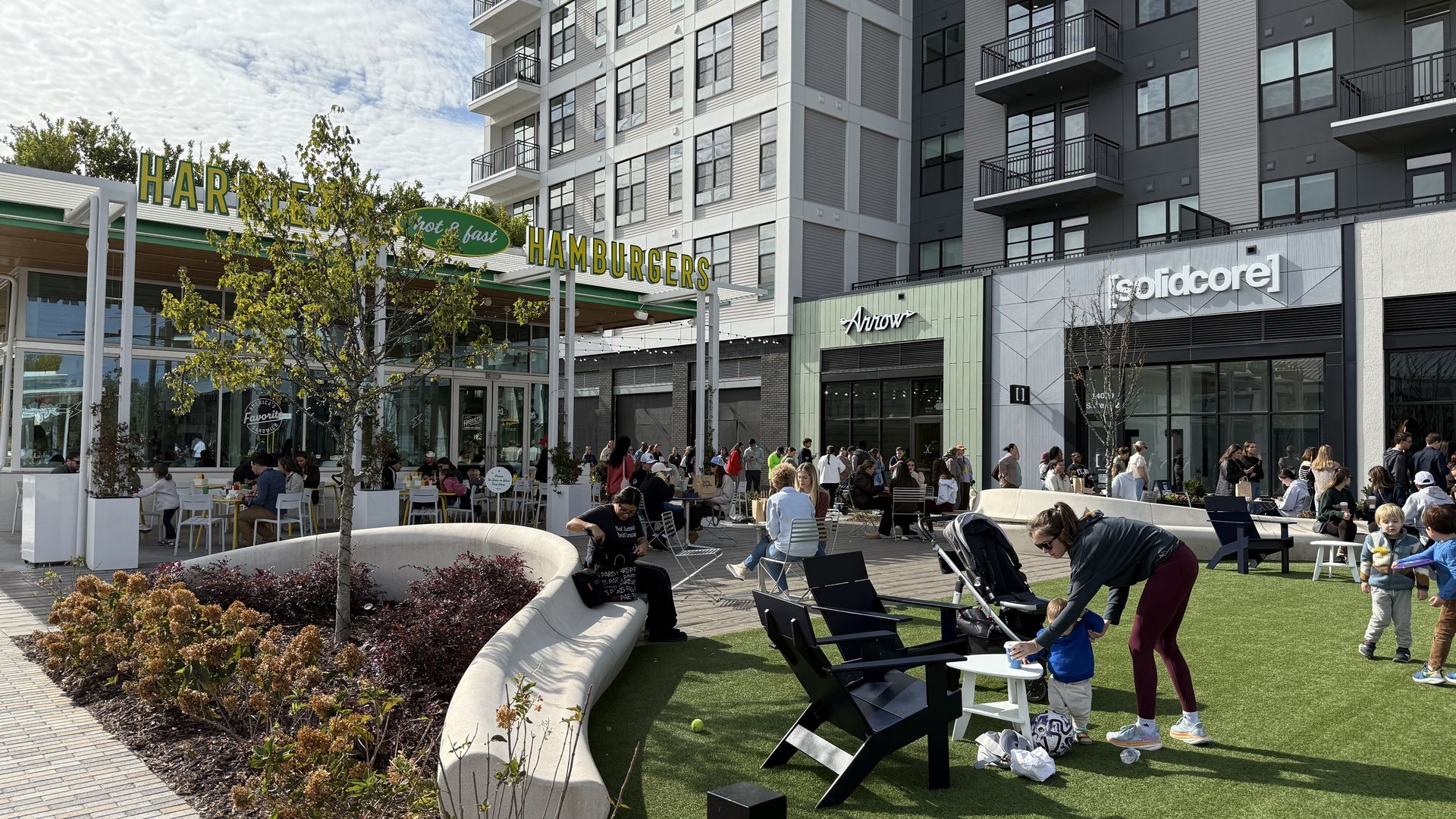 Outdoor urban plaza with people sitting and dining near green artificial turf, a curved concrete bench, black chairs, and storefronts including a hamburger restaurant, Arrow, and solidcore.