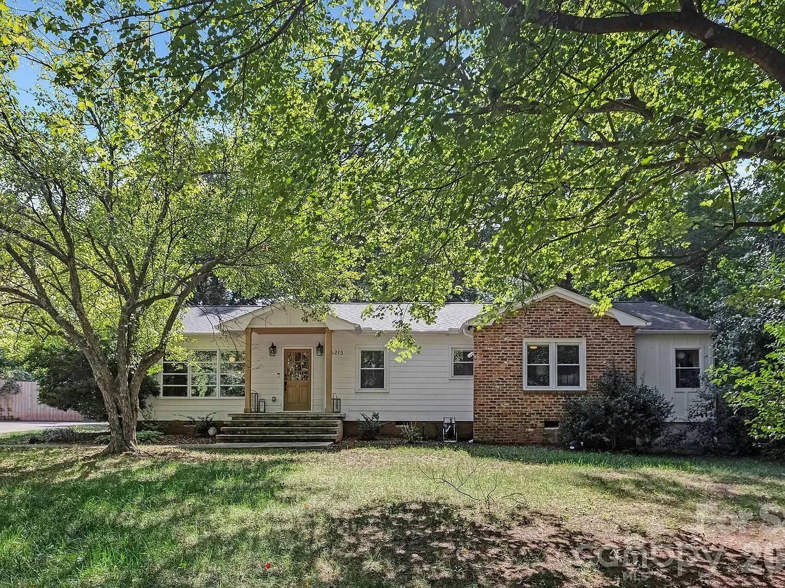 Front view of a one-story house with white siding and a brick chimney on the right. A wooden front door with a small porch, steps, and green lawn; large tree branches shade the yard.