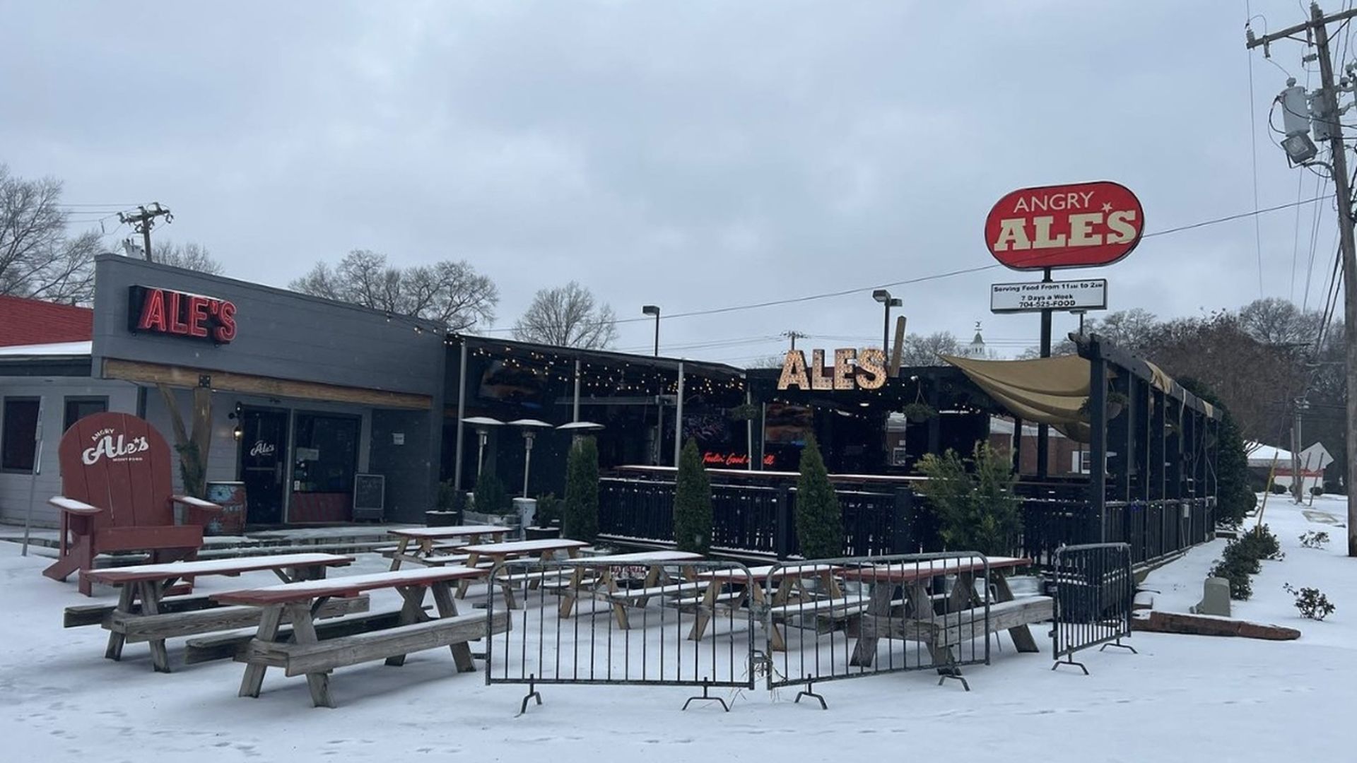 Snow-covered outdoor seating area with picnic tables and black fences in front of Angry Ale's bar and grill under a cloudy sky.