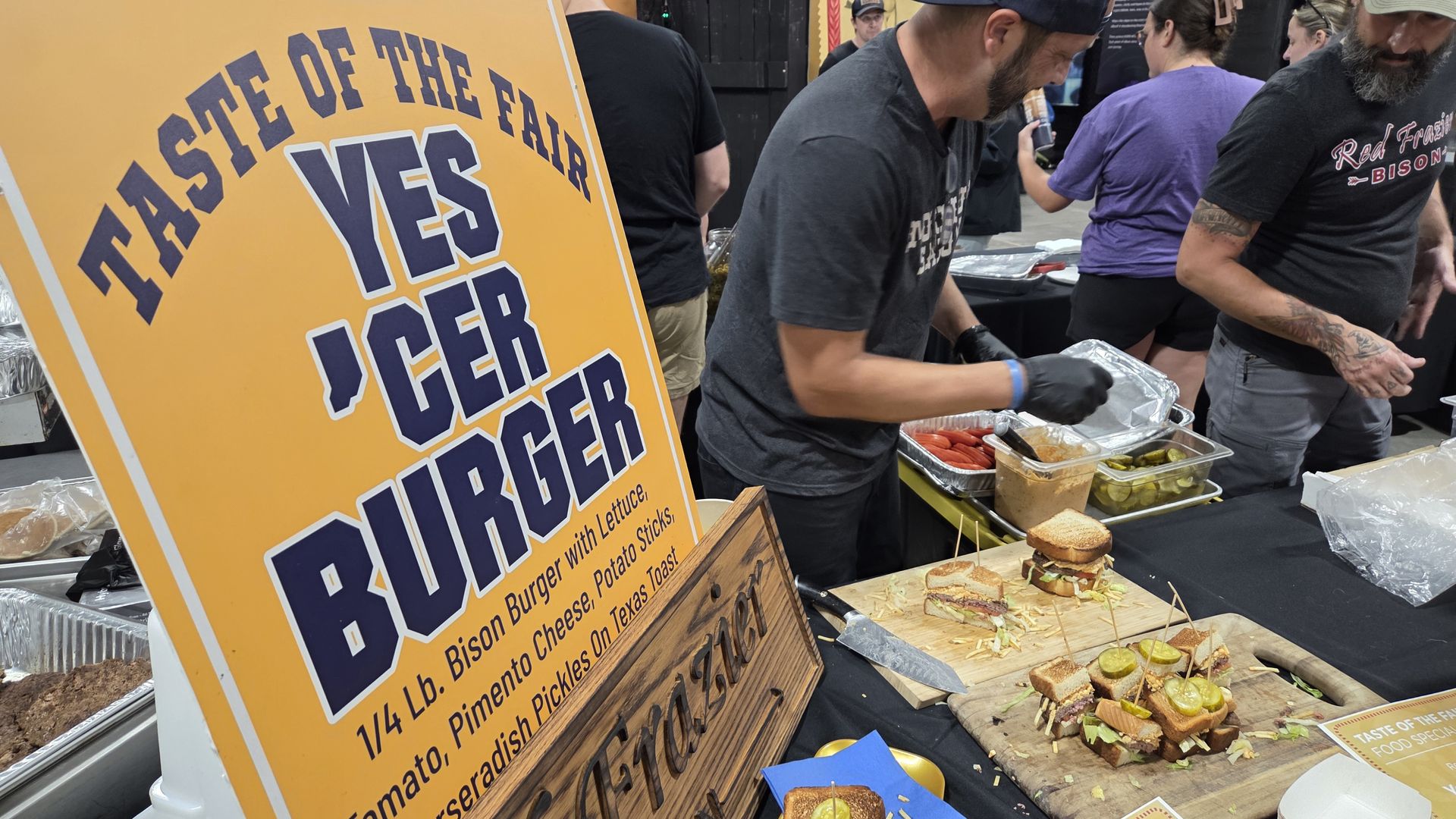 People preparing and serving bison burgers at a fair booth with a large yellow sign reading "Taste of the Fair YES 'CER BURGER" describing a 1/4 lb bison burger with toppings on Texas toast.