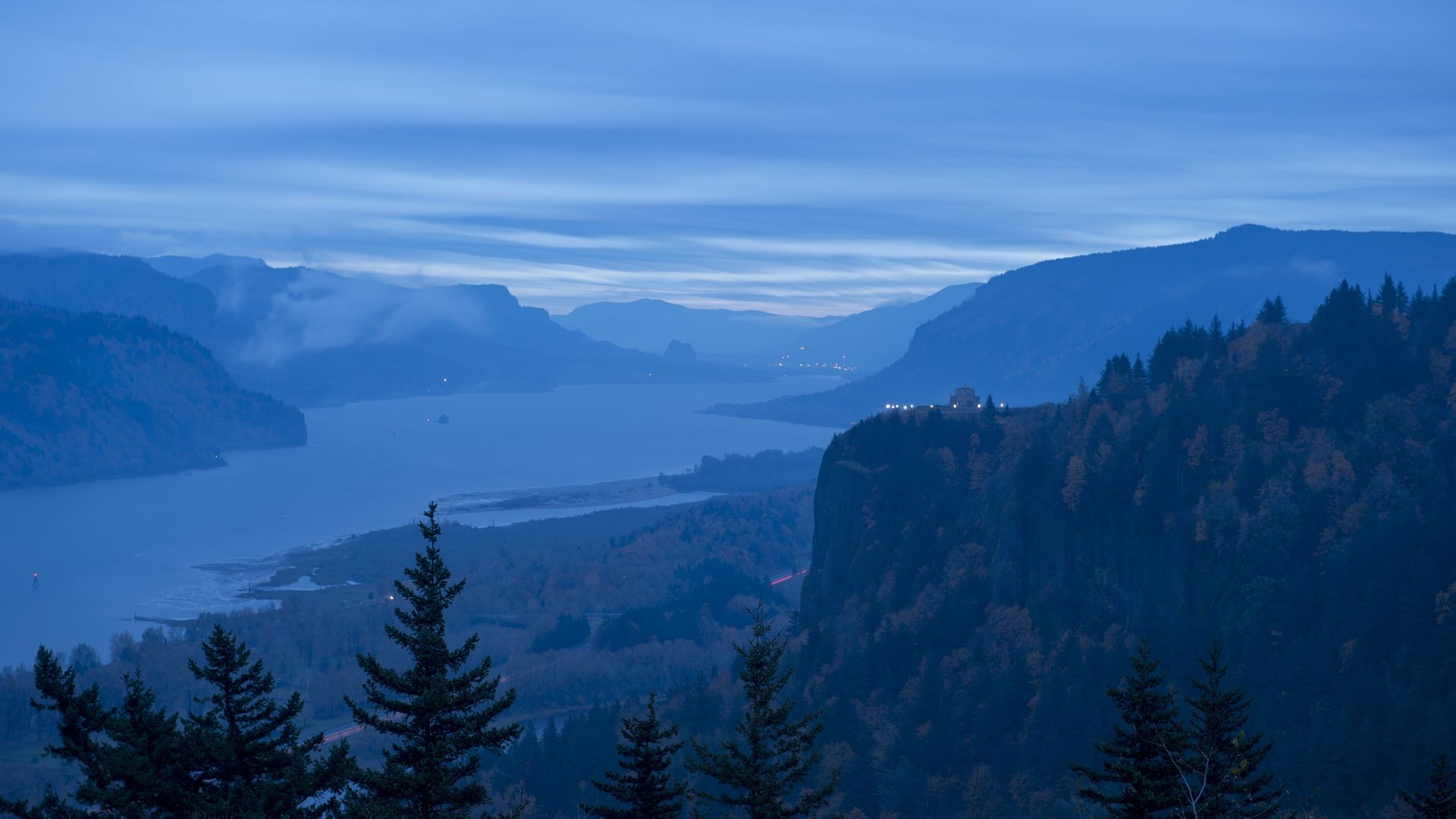 Blue-tinted twilight view of a river winding through forested mountains, with a cliff topped by a small lit building on the right and silhouetted pine trees in the foreground.