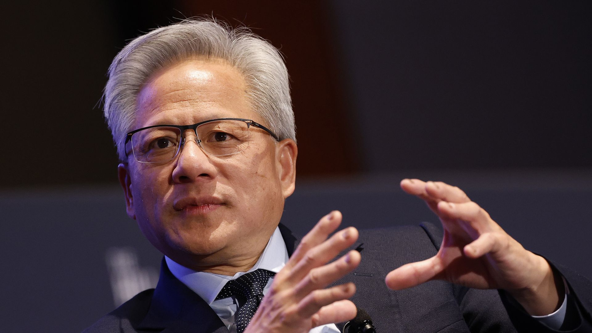 Close-up of a man with gray hair and glasses, wearing a dark suit and patterned tie, speaking while gesturing with his hands.