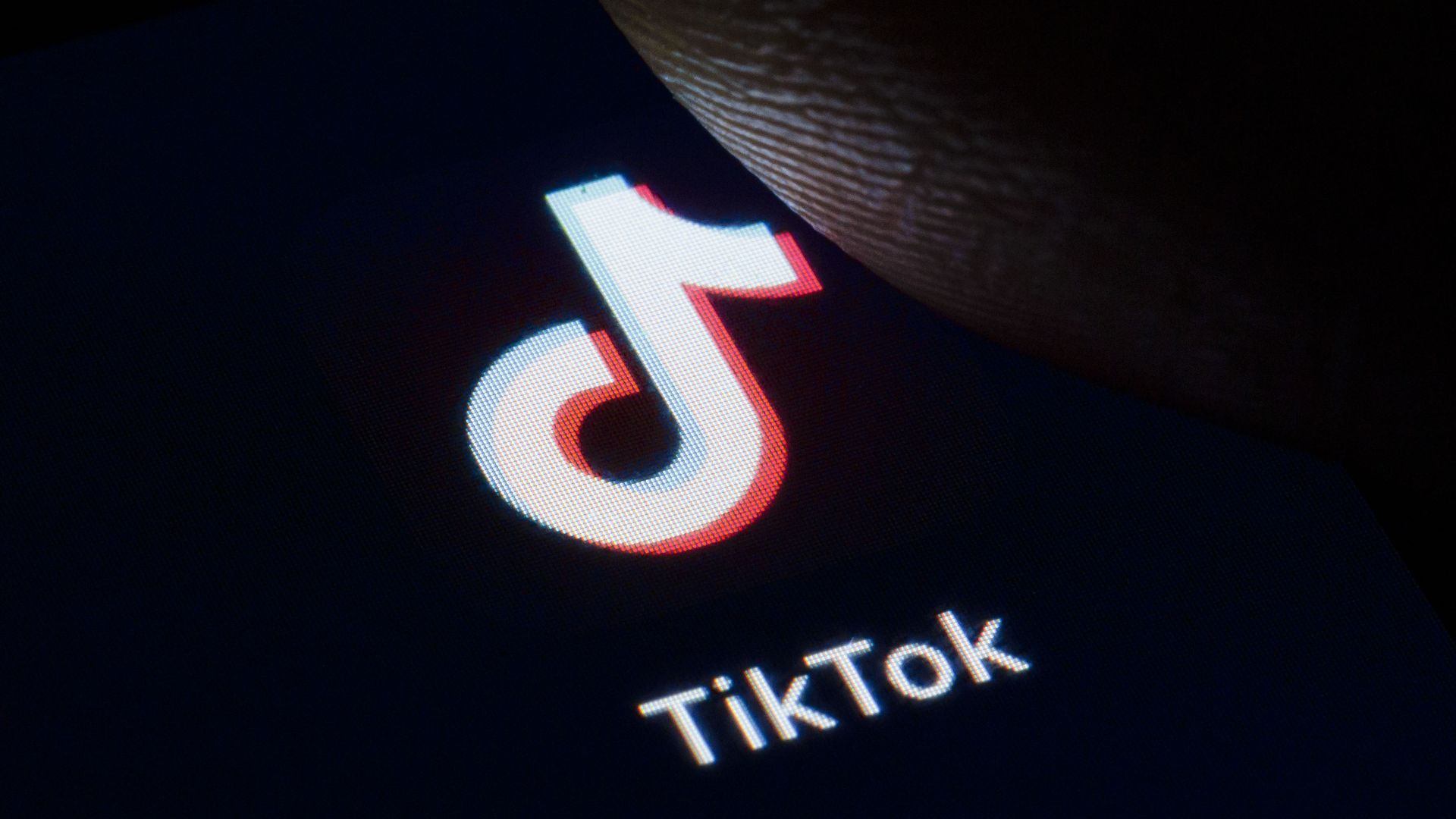 In this image, a thumb hovers over the TikTok logo displayed on a smartphone screen.