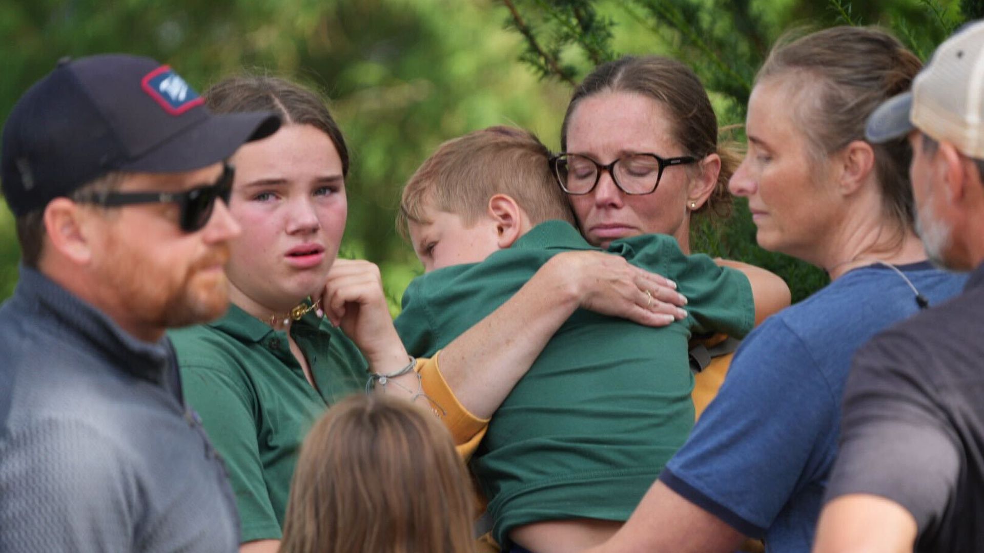 A group of people outdoors share a somber moment; a woman with glasses hugs a young boy in green while another girl in green looks upset, surrounded by others with serious expressions.