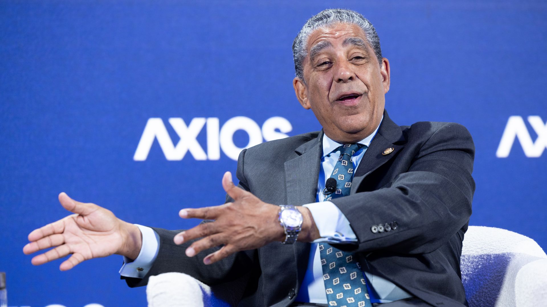 An older man in a dark suit and patterned tie sits in a white chair on a blue backdrop, gesturing with his hands while speaking; the word "Axios" is visible behind him.