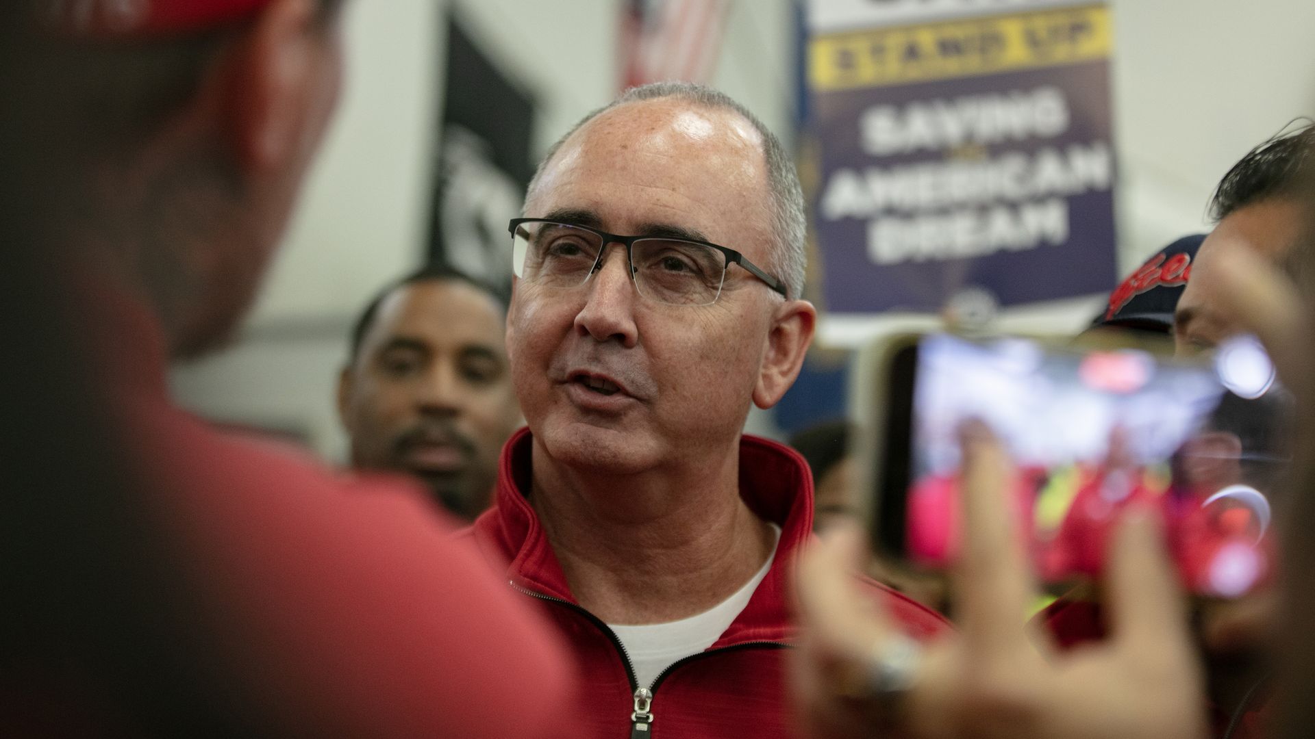 A person in glasses speaks to supporters in a room with a union picket sign being held up in the background