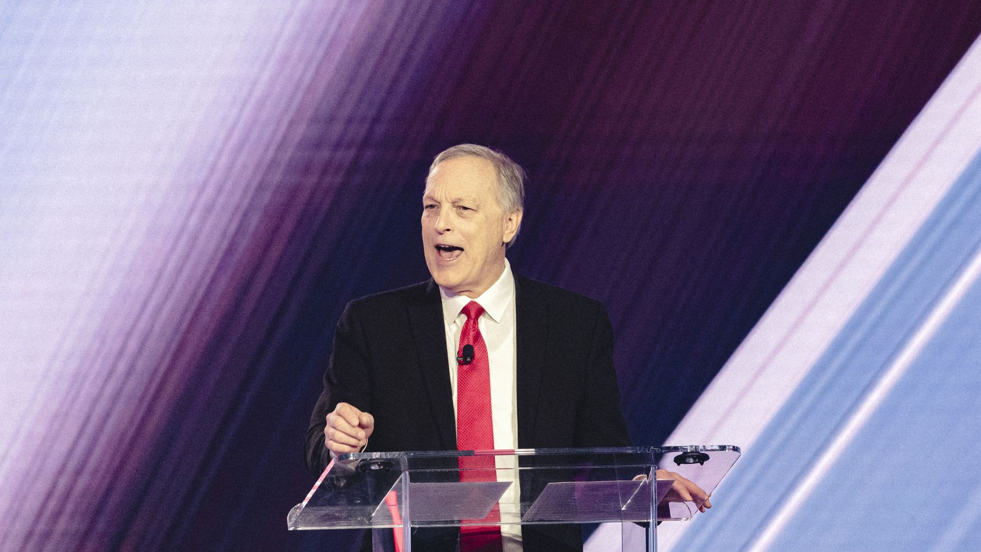 A man in a black suit, white shirt and red tie speaks at a clear lectern, in front of a multicolored background with broad streaks of purple and blue.