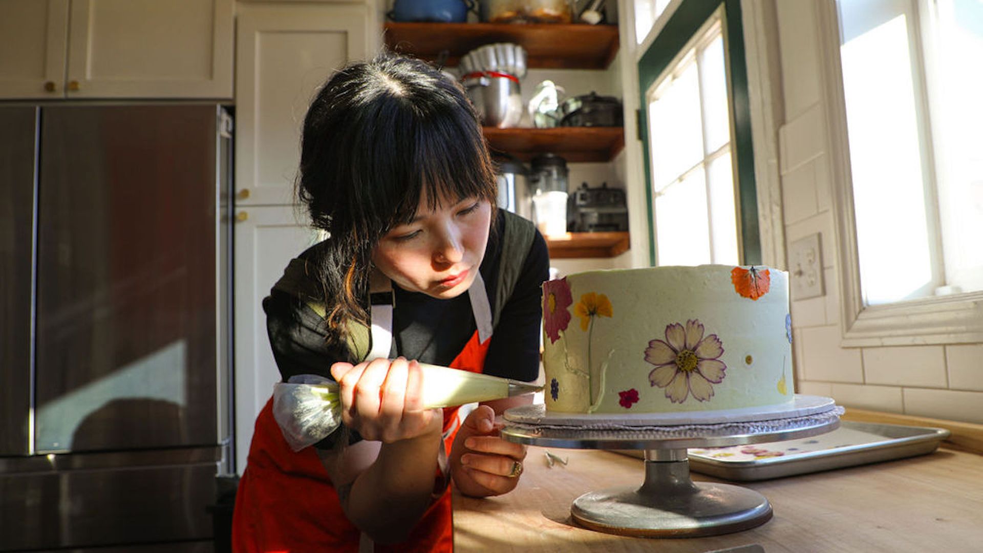 A woman in a red apron decorates a round cake with pastel floral designs using a piping bag in a bright kitchen, cake on a stand near a sunlit window.