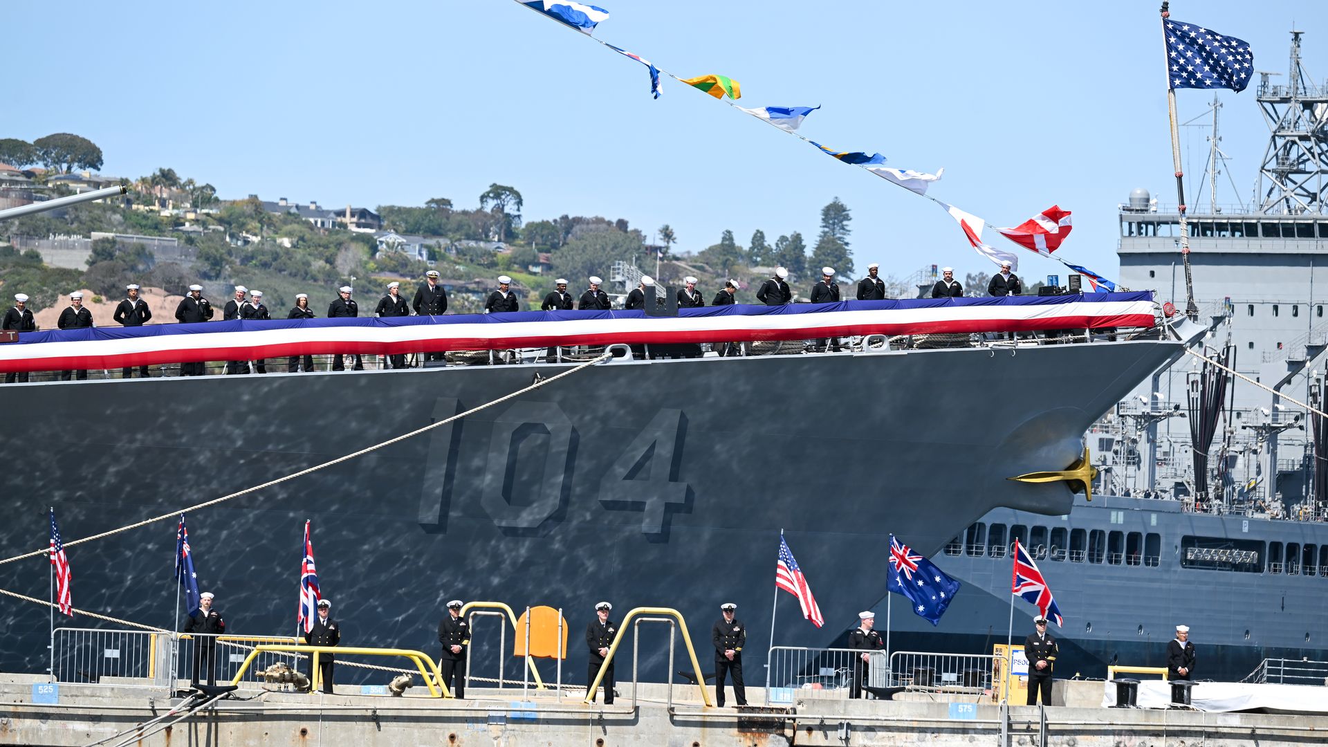 Marines and sailors in uniform stand on a navy ship with different country's flags flying and on a platform below the ship at Naval Base Point Loma in San Diego.