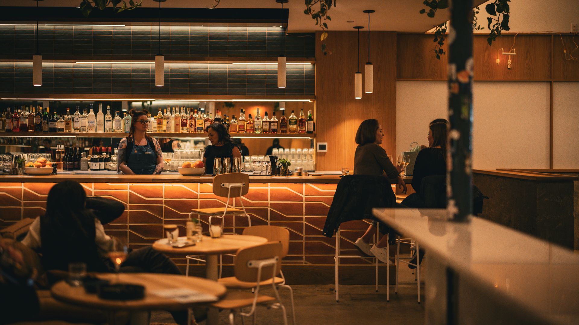 A dimly lit bar with two bartenders behind a bar stand, while two other people chat while seated nearby. There is a row of liquor bottles on shelves behind the bar. In the foreground, several coffee tables and chairs are visible. 