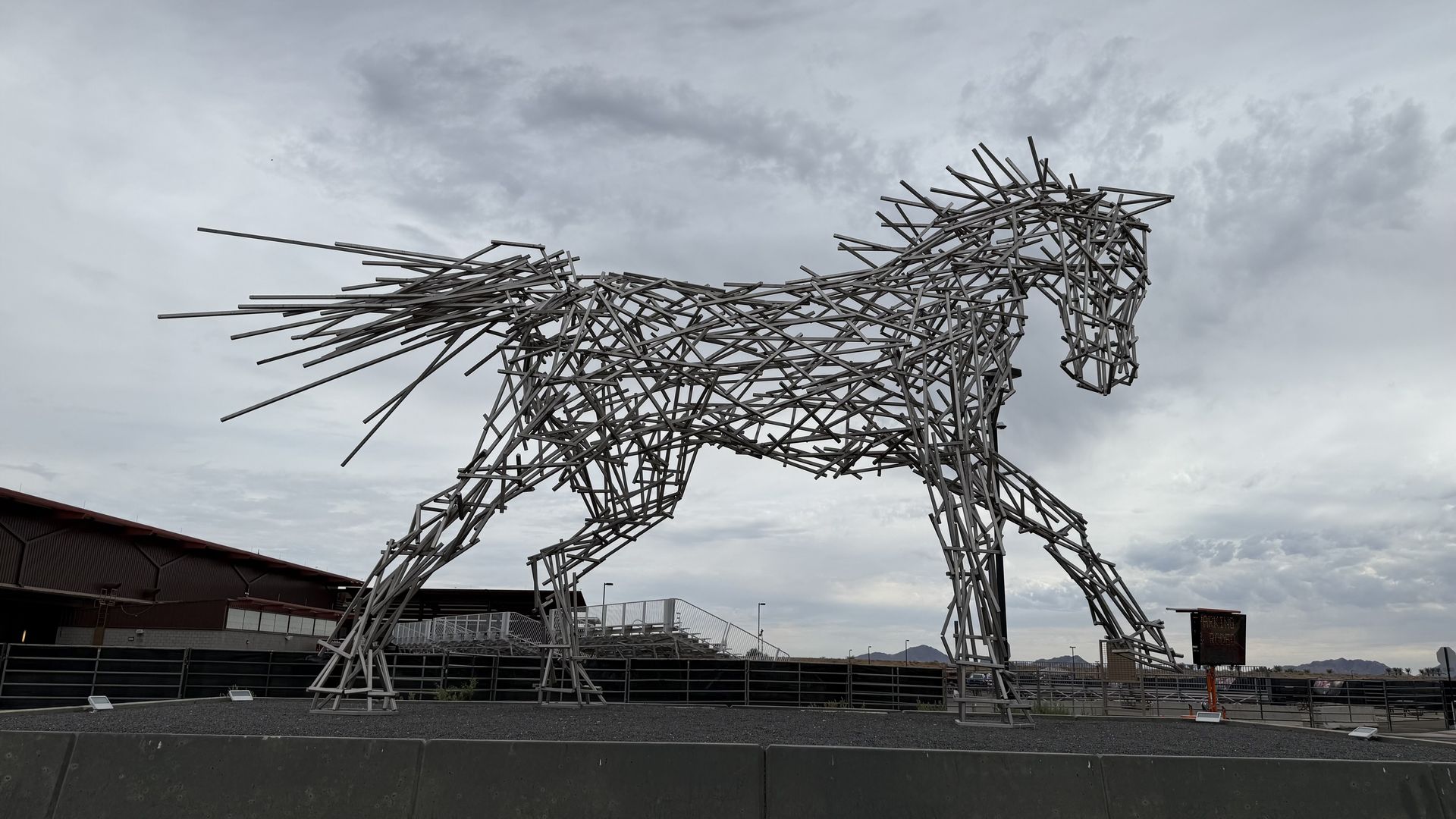 Outdoor sculpture of a horse formed from a lattice of steel rods, standing against a cloudy sky in a plaza with fences and distant mountains.
