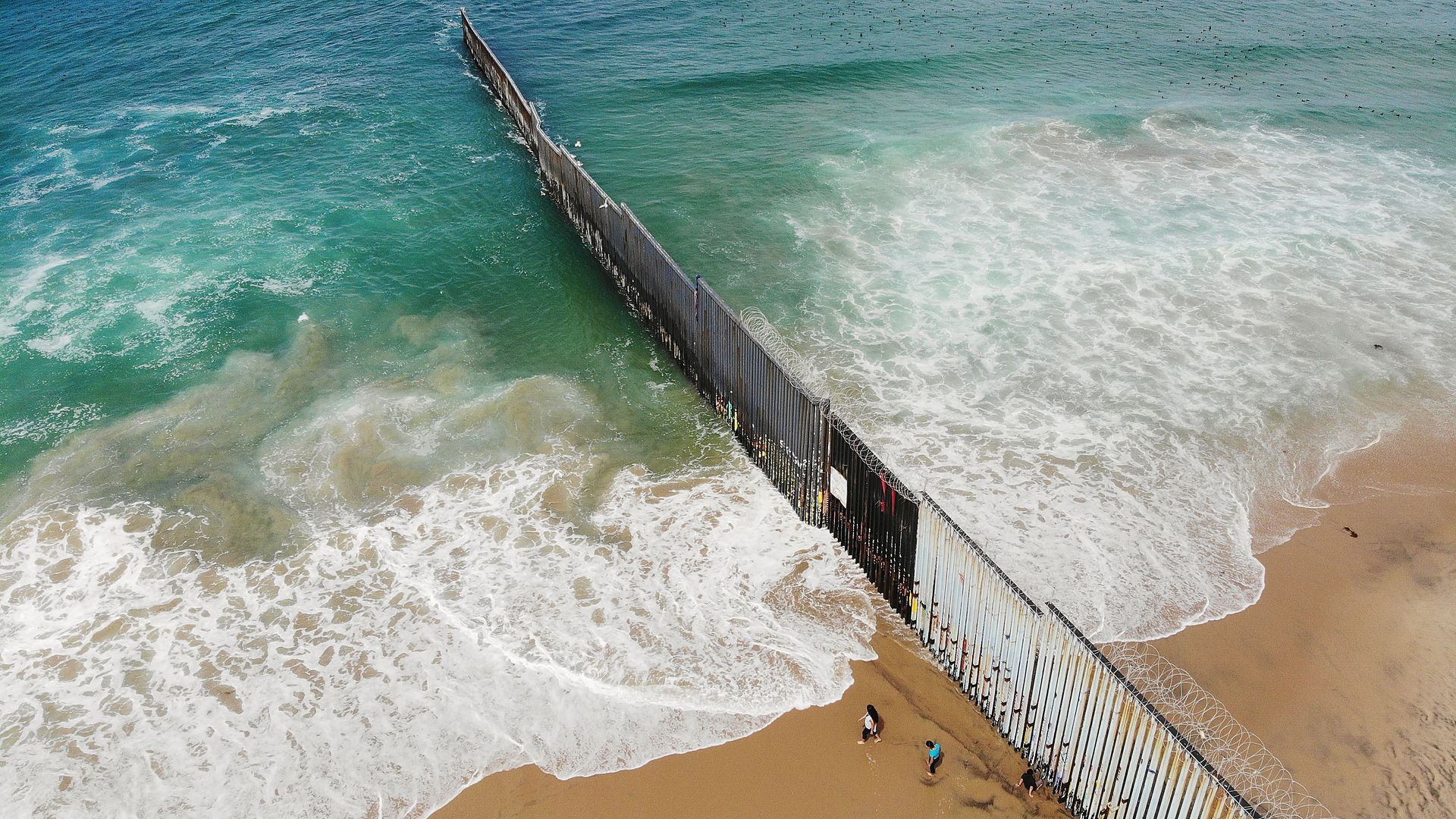 In this image, three people from Mexico walk along a tall fence the southern border that leads down the beach and into the ocean.