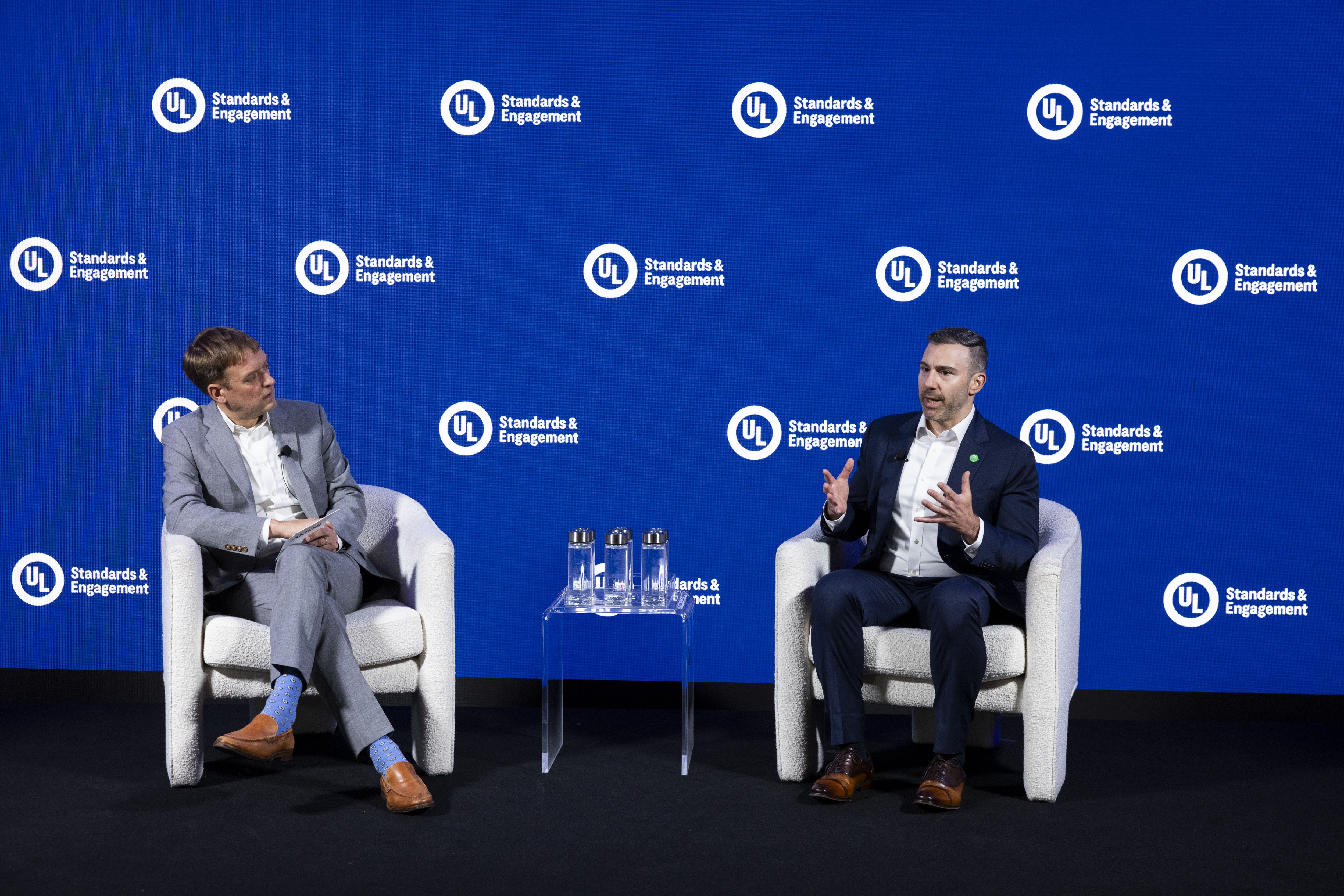 Two men in suits sit in white chairs on a stage with a blue backdrop featuring repeated UL Standards & Engagement logos; a clear table with water bottles sits between them as they talk.