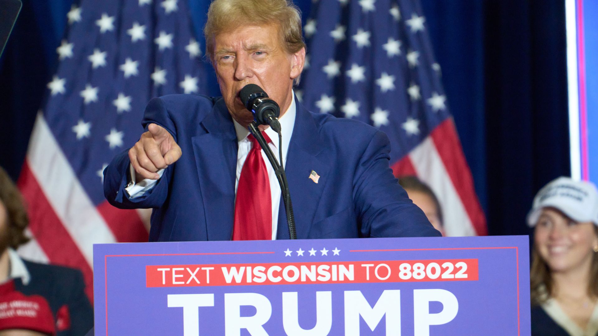 Former President Trump, wearing a blue suit, white shirt and red tie, speaks at a rally in front of supporters and American flags.