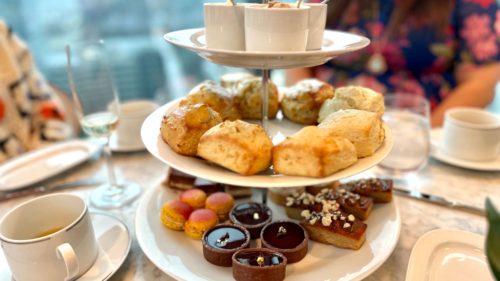 Two-tiered white serving tray with scones on the top plate, chocolate tarts, macarons, and nut-topped bars on the bottom, surrounded by teacups and glasses on a marble table.