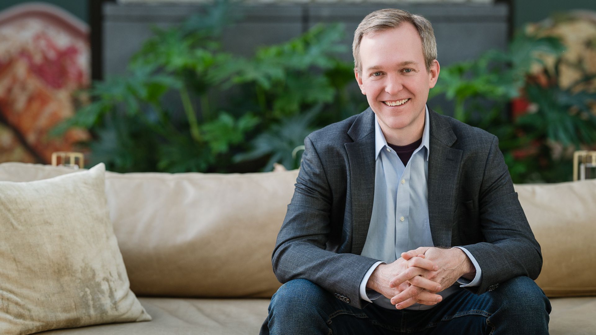 Smiling man in a blazer sits on a beige sofa with folded hands, wearing a light blue shirt and jeans; leafy green plants and a modern indoor backdrop surround him.