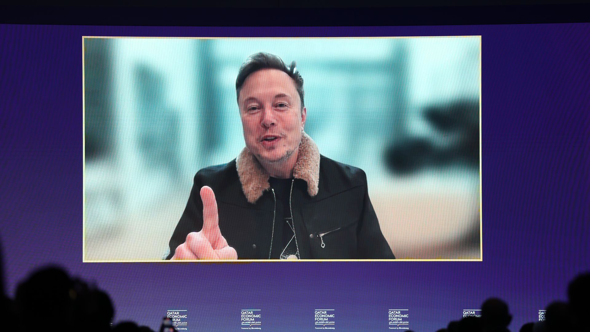 A person in a jacket with a wool collar points at a camera while speaking into it