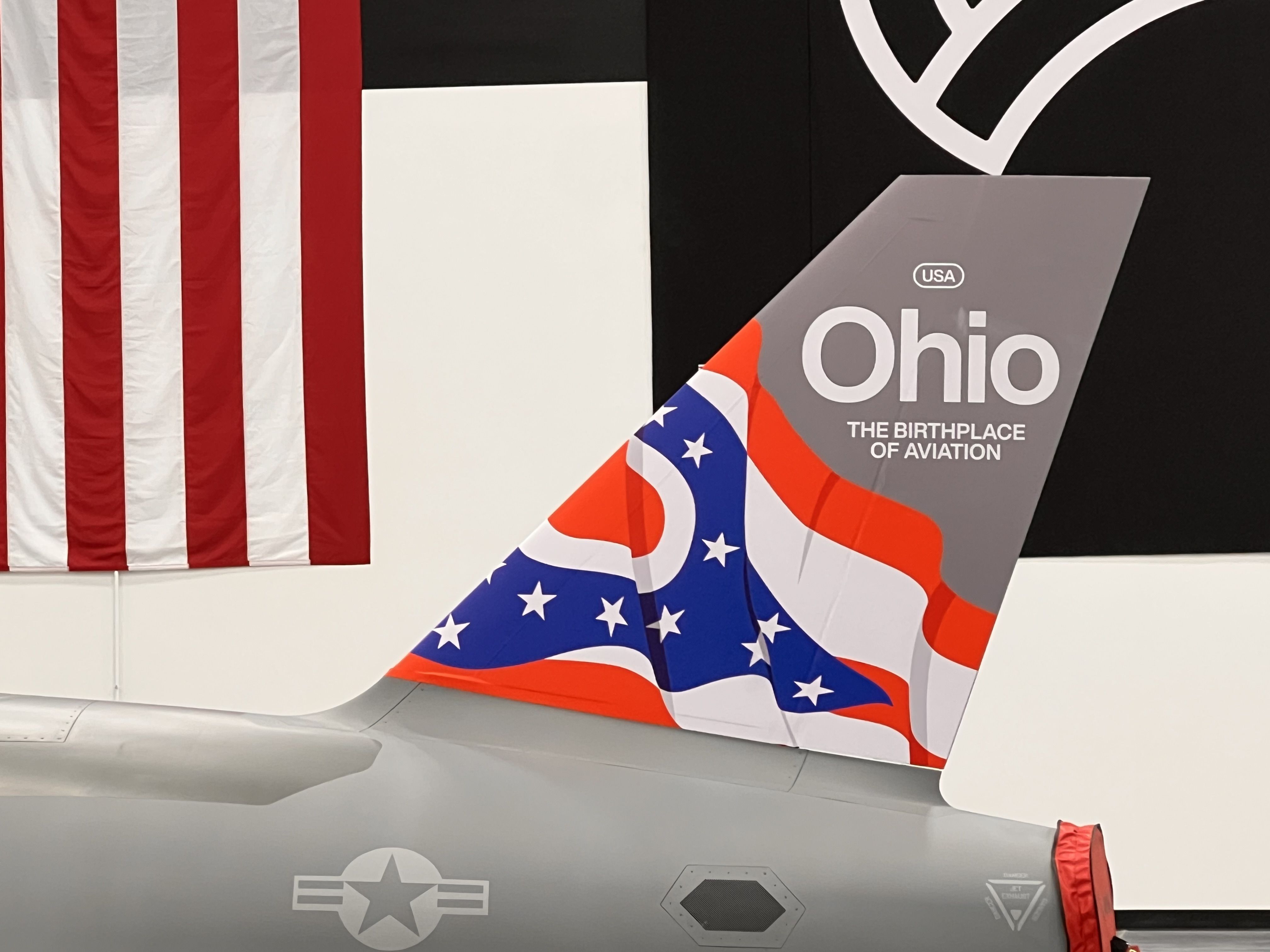 Exhibit scene: left shows a tall American flag; right features a gray aircraft tail with USA and Ohio and the title "The Birthplace of Aviation" on a black wall; red, white, and blue fabric drapes over the tail.