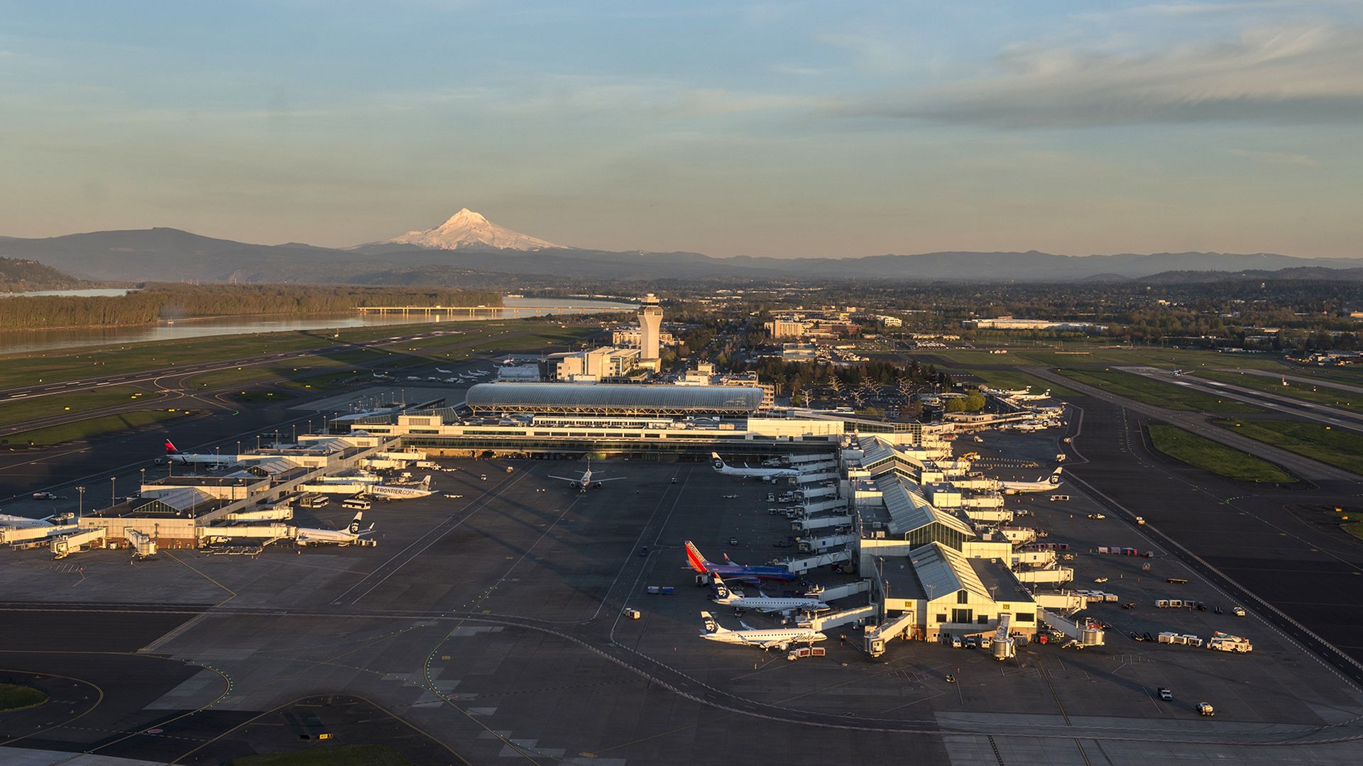 An aerial shot shows airplanes parked at the Portland International Airport with Mount Hood looming in the background.