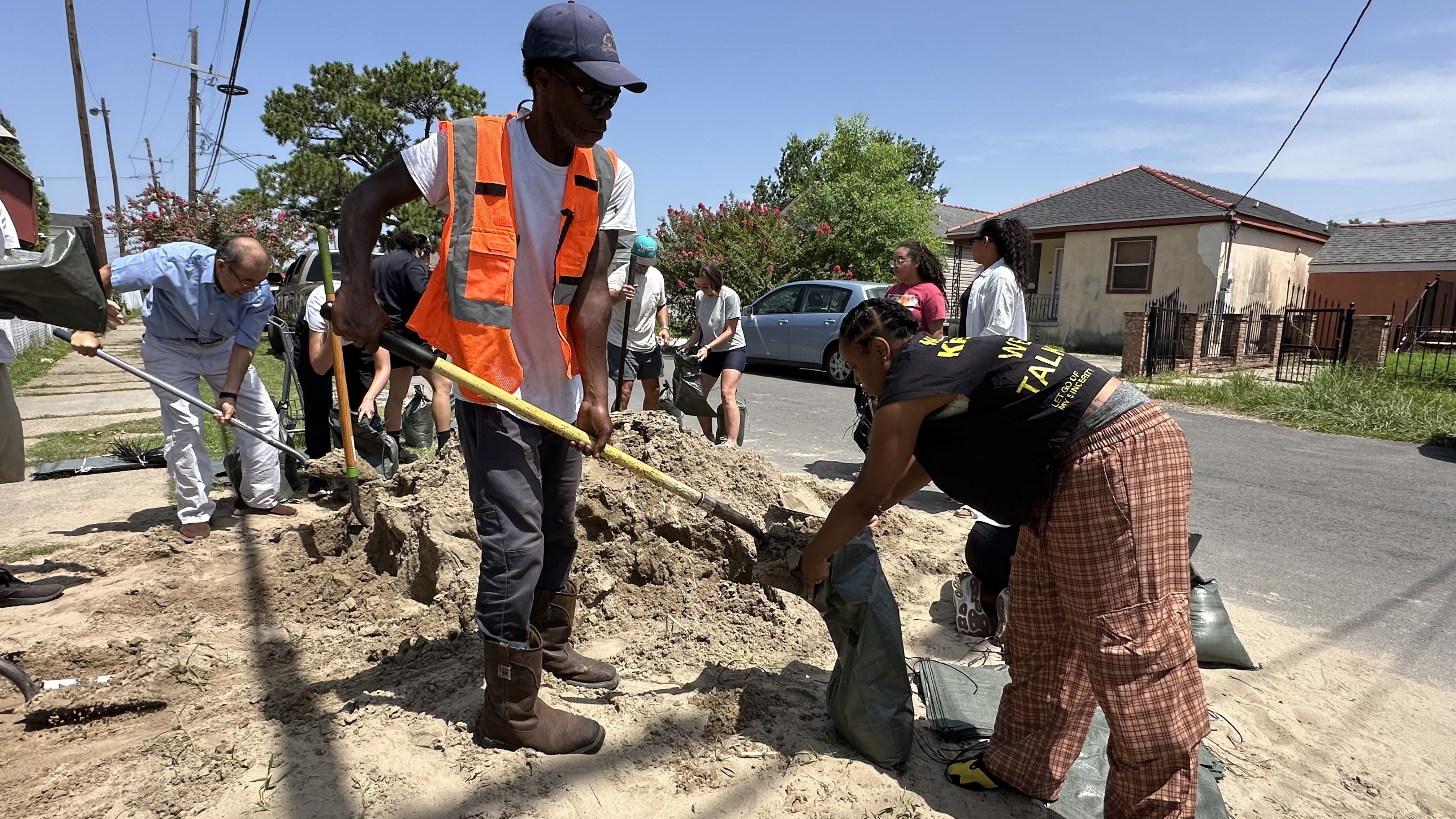 Group of volunteers in orange safety vests use shovels to dig a large dirt pile on a sunny suburban street; bags, a parked car, and houses are visible in the background.
