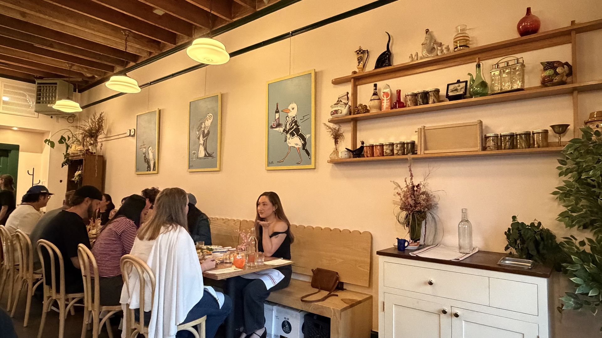 Cozy restaurant with exposed wooden beams and warm pendant lights. Long table of diners sits under blue cartoon prints on the wall, with wooden shelves holding bottles, jars, and knickknacks; white cabinet to the right.