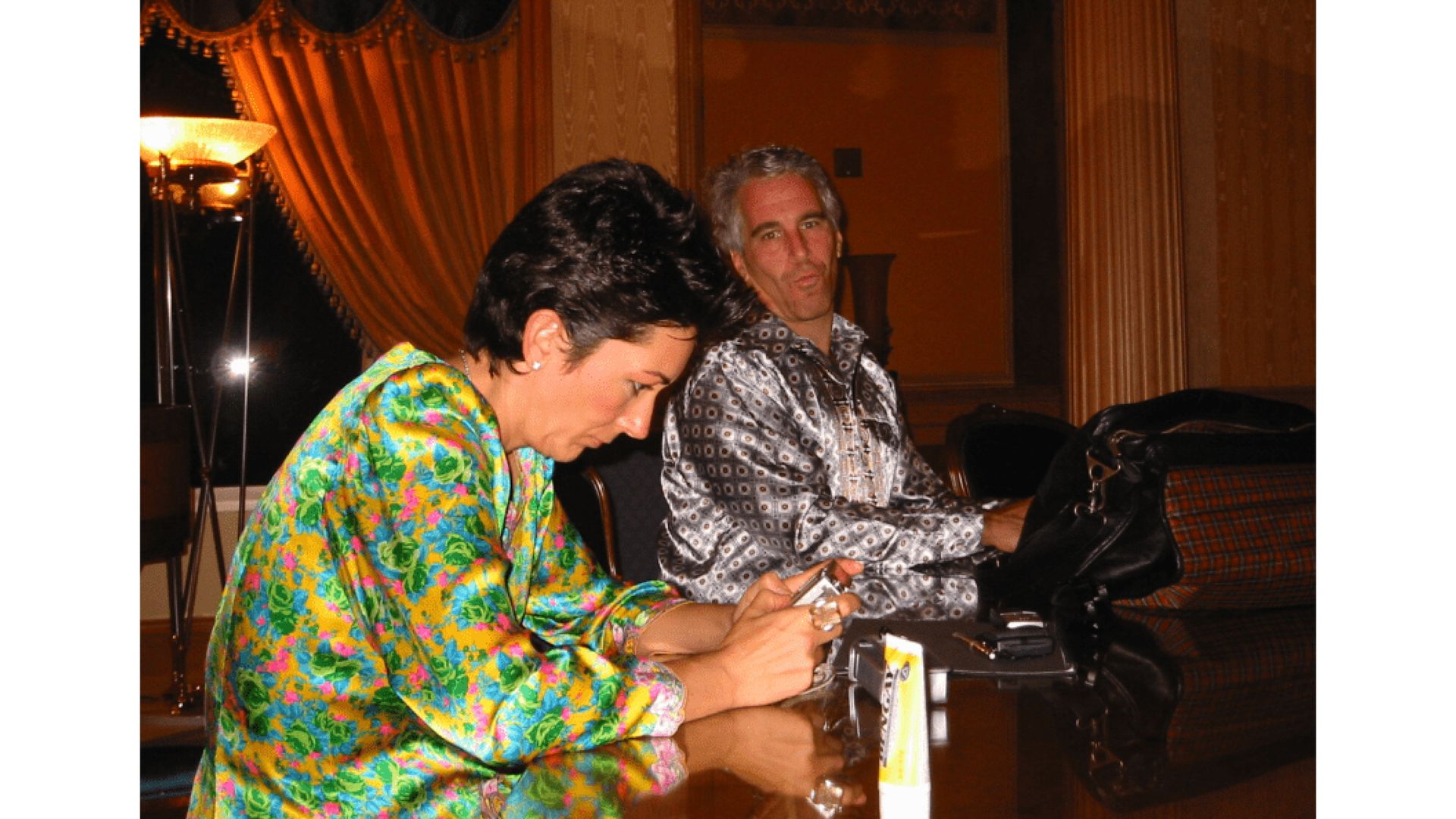 Two people sitting at a table in a warmly lit room with gold curtains. The person on the left wears a bright floral jacket and looks at a device; the person on the right wears a patterned shirt.