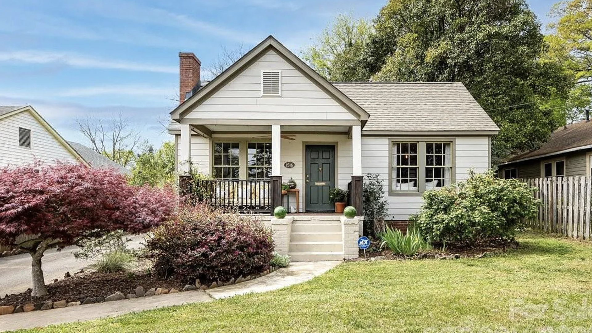 Cozy, single-story white cottage with a gray gabled roof, green front door, and small porch. Red-leaf shrub left, green bushes right, blue sky, brick chimney, and a fenced yard.
