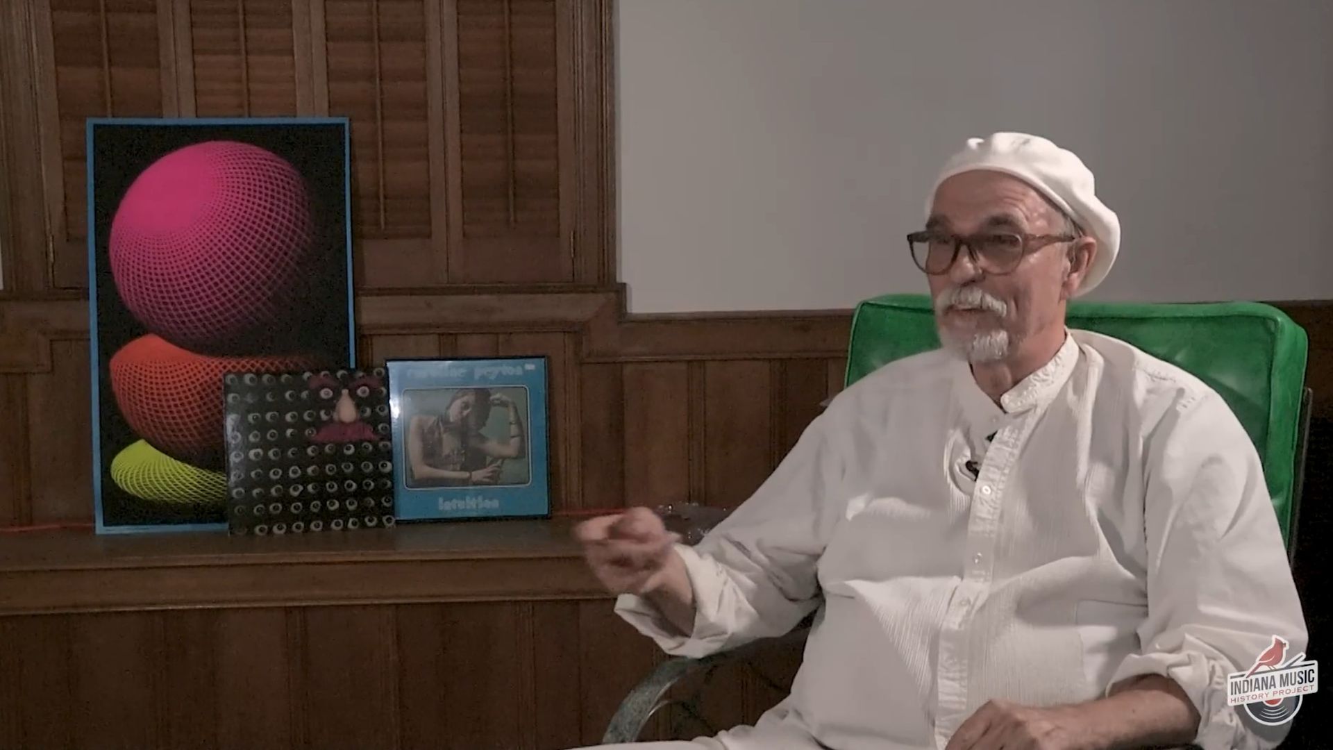 Elderly man in white shirt and beret sitting on a green chair gesturing with hand, three colorful album covers on wooden shelf behind him, Indiana Music History Project logo at bottom right.