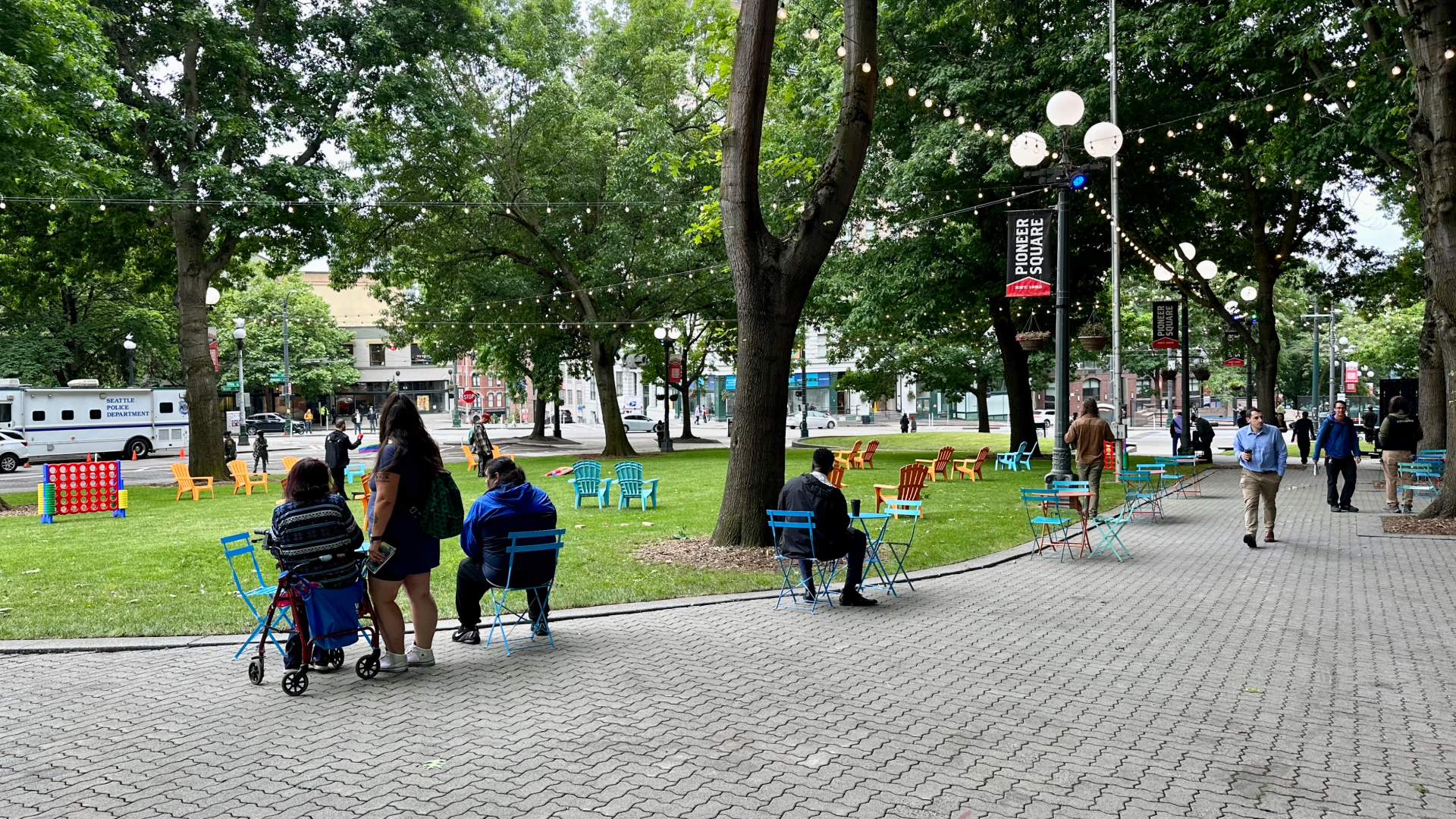 People sit at cafe tables on a paved area that abuts a grassy area, with lights strung overhead and games set up on the lawn. 