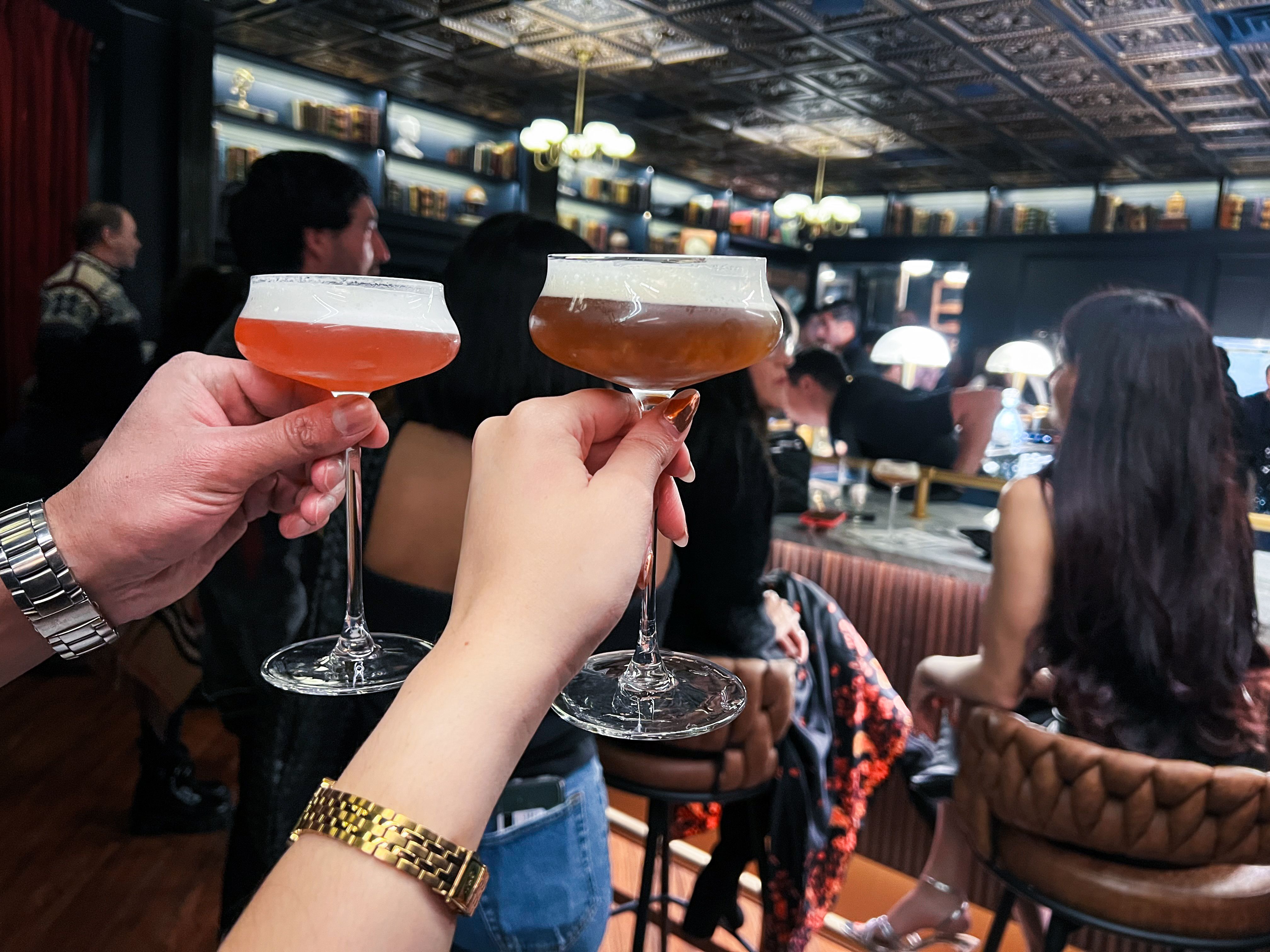 Two hands holding long-stemmed cocktail glasses with brown-hued drinks. 