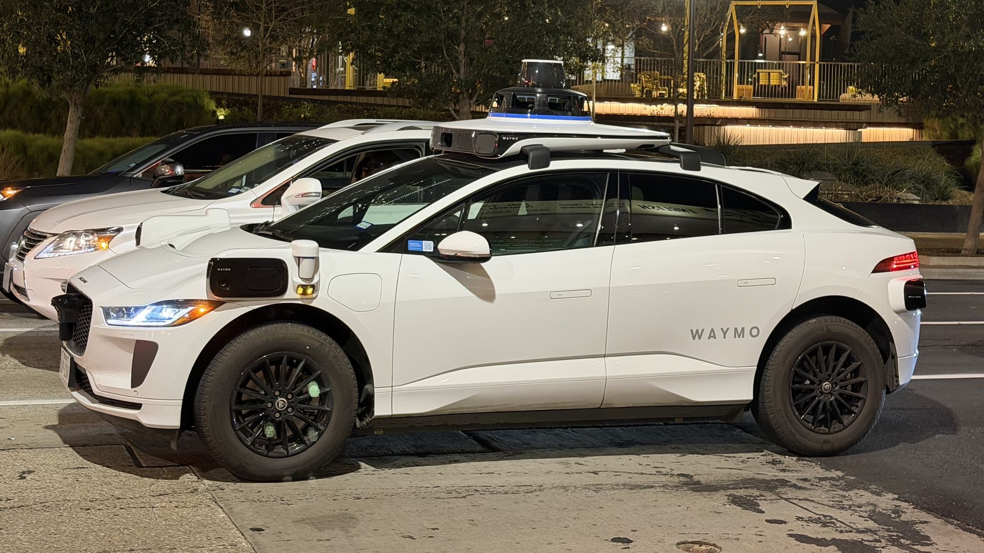 Nighttime parking lot with white Waymo self-driving cars in the foreground, bright streetlights, trees, a yellow playground frame behind a fence, and tall buildings in the background.