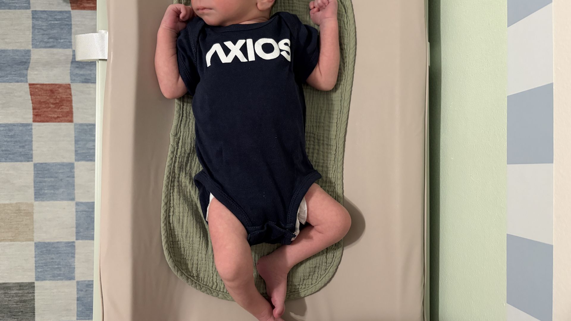 Baby on a green quilted changing mat on a beige surface, wearing a navy onesie that reads "AXIOS"; arms near head, legs crossed. Left shows blue checkered tiles, right shows pastel stripes.