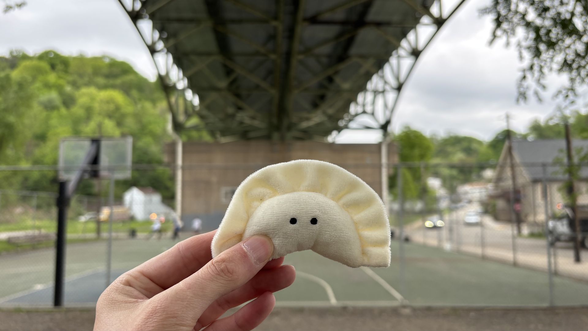 Close-up of a cream-colored, dumpling-shaped plush with two black eyes held by a hand, outdoors on a tennis court with a large metal overhead structure and green trees in the background.