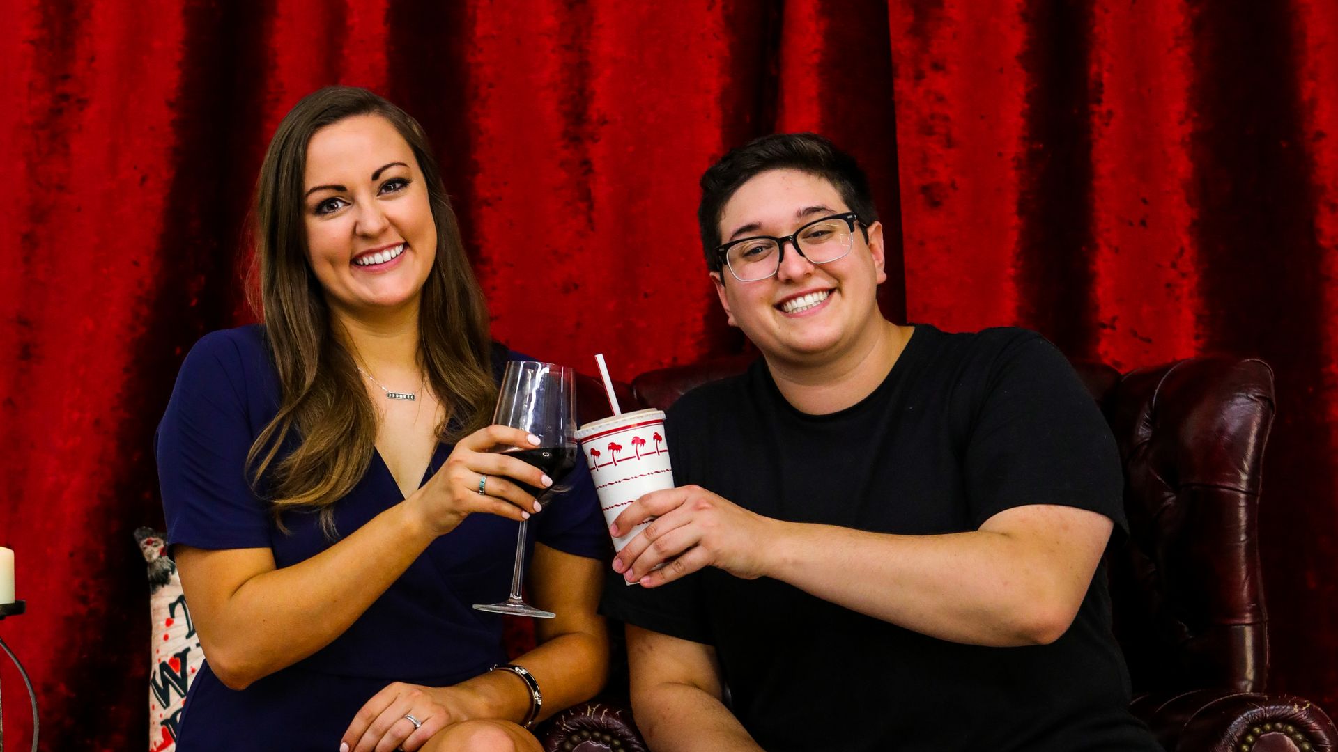 Two women cheers each other, one with a wine glass, the other with a fast food beverage. 