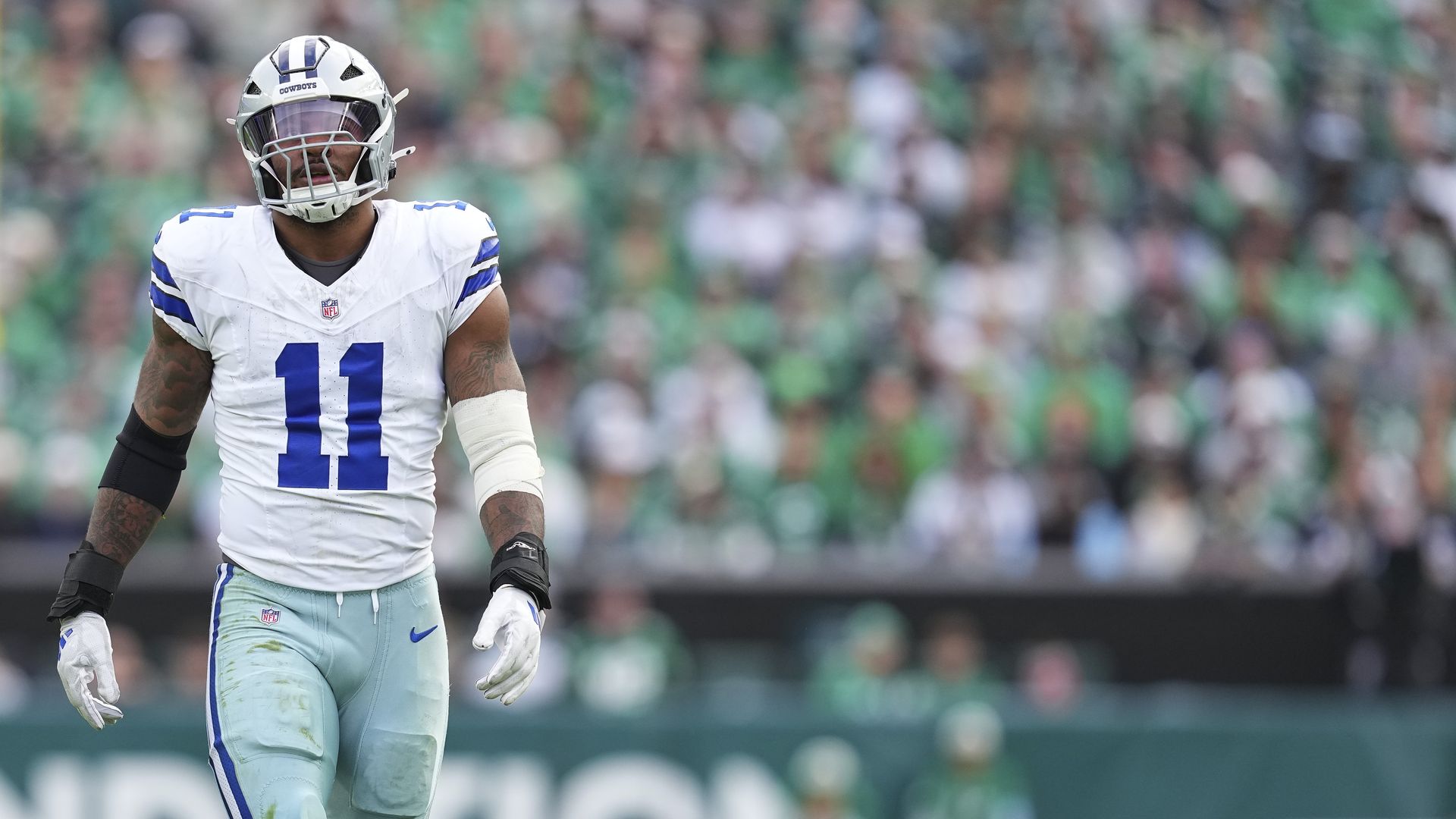 Dallas Cowboys football player in white and blue uniform with number 11 on field, standing with posture, blurred crowd in green in background.