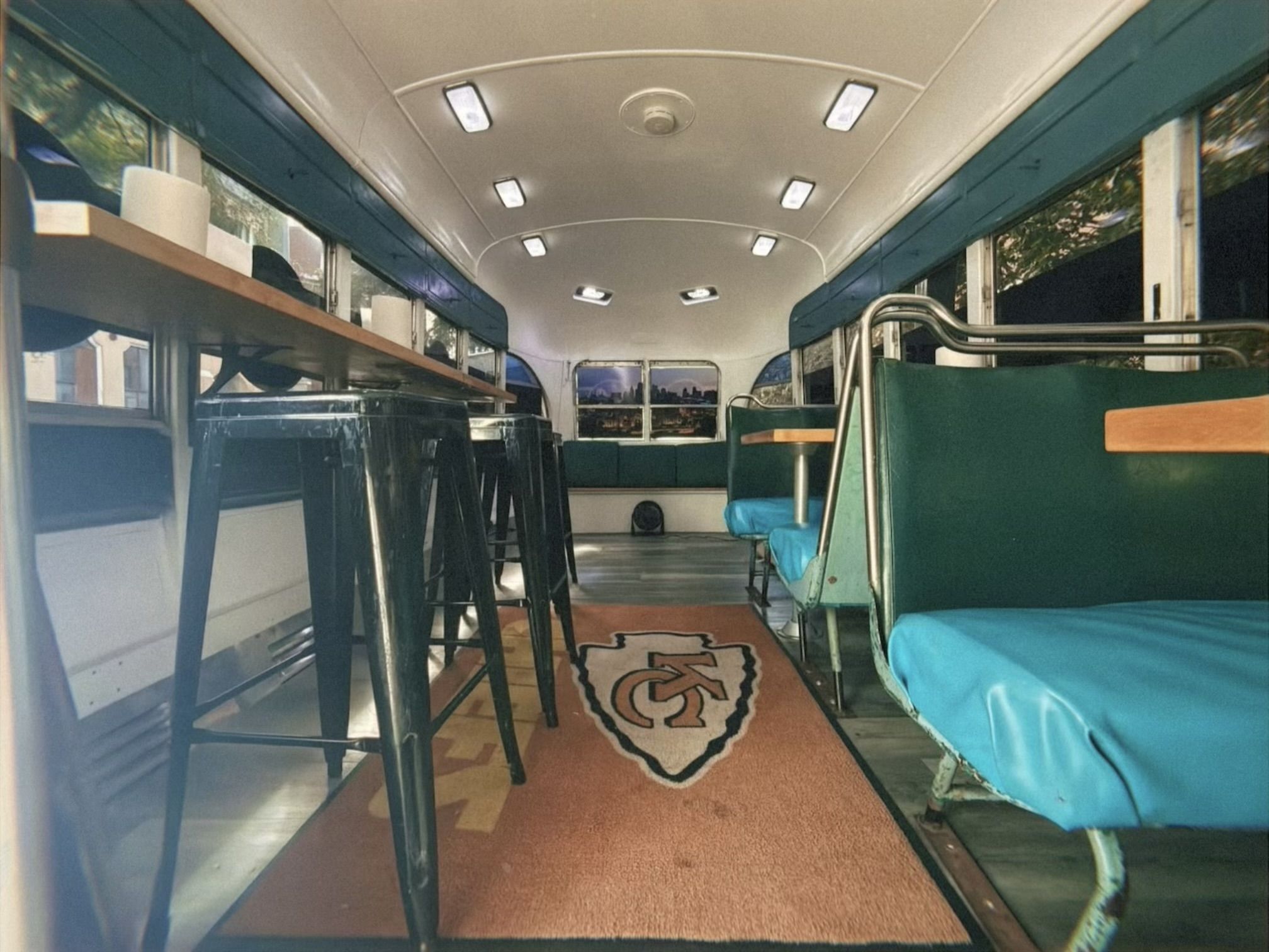 Interior of a bus converted into a lounge with teal bench seating, metal stools, a long wooden shelf, and a red rug featuring the Kansas City Chiefs logo.
