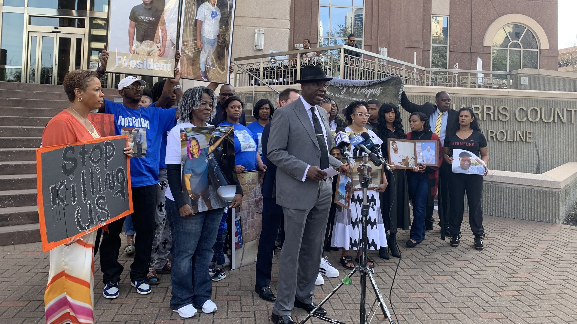 Photo of a press conference with a man in a suit speaking in the front and people holding photos in the back.