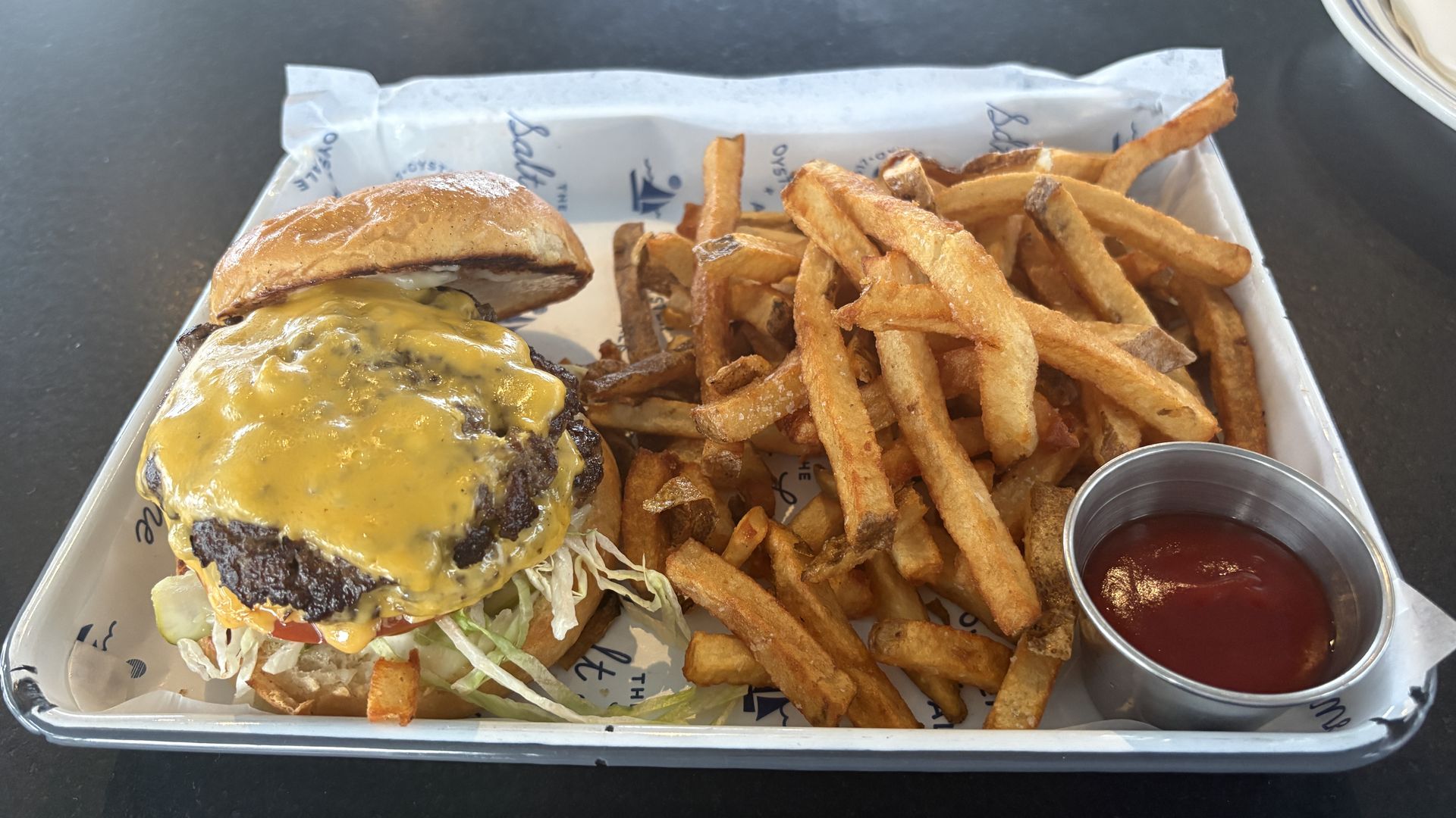 Cheeseburger with melted cheddar on a beef patty in a toasted bun, with lettuce and tomato, beside a mound of crispy fries and a cup of ketchup on a metal tray.
