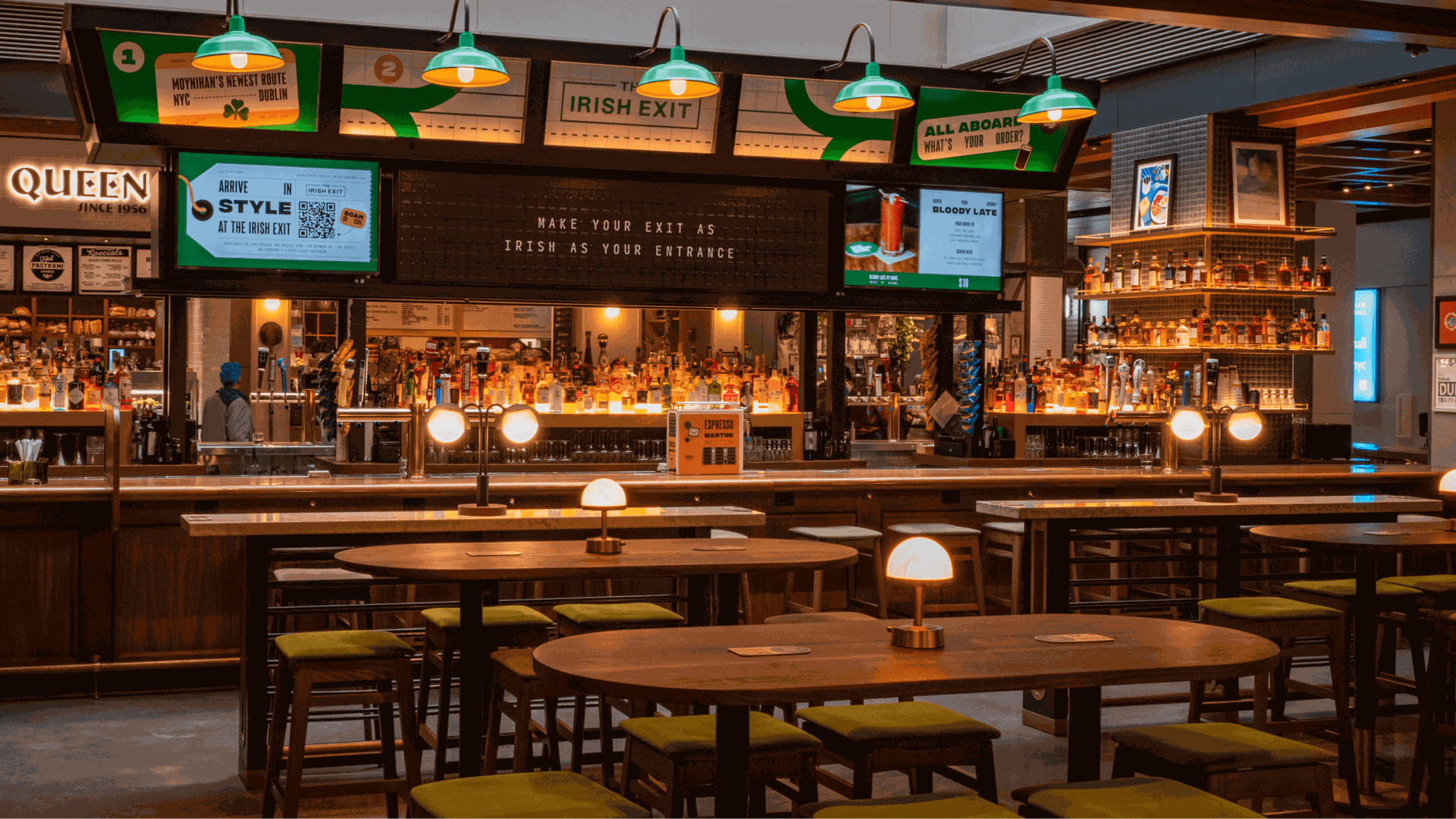 Warmly lit bar interior with wooden tables, green cushioned stools, and a backlit shelf of bottles. Green pendant lamps hang above, signs read "The Irish Exit" and drink menus.