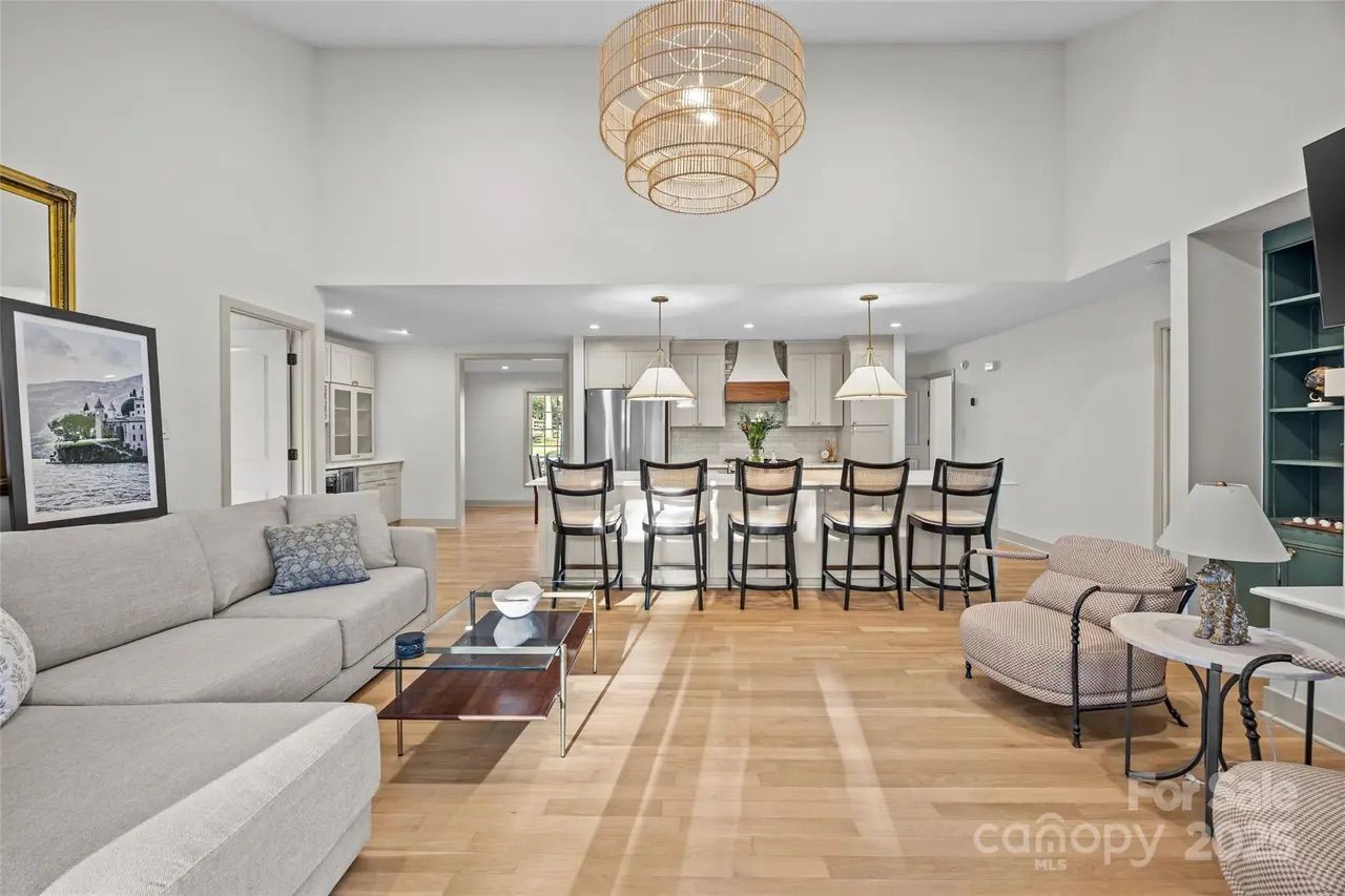 Open living space with light wood flooring, beige sectional sofa, glass coffee table, kitchen island with five dark chairs, white cabinetry, and a large woven ceiling light fixture.