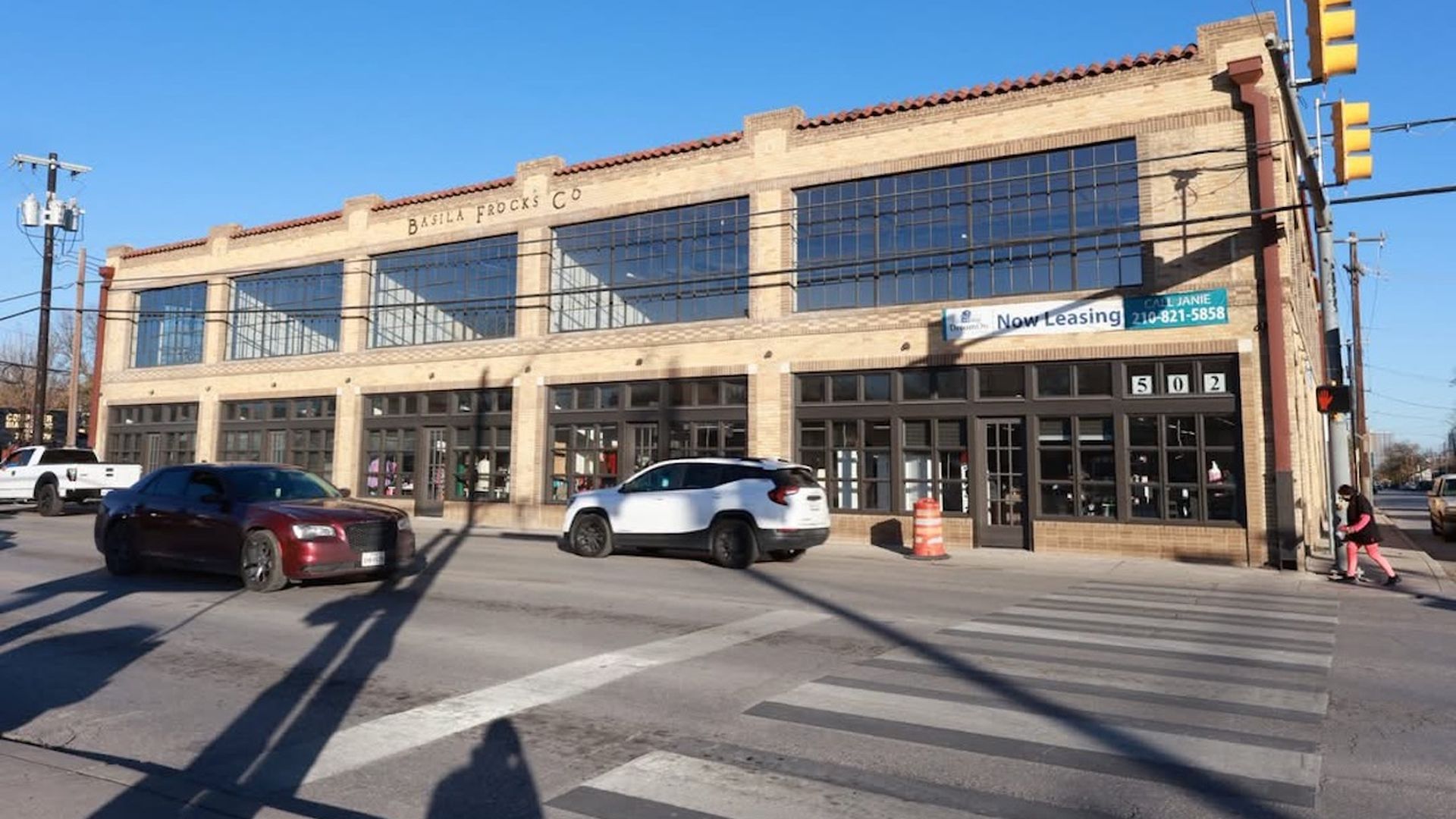 Two-story brick storefront building with large windows, Basilia Frocks Co sign, on a street with crosswalk, yellow traffic lights, parked cars, and a clear blue sky.