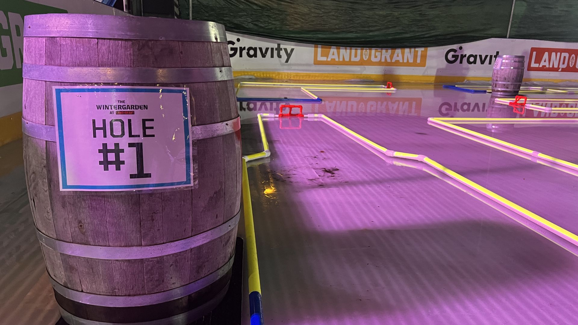 Wooden barrel marked "Hole #1" next to neon-bordered mini golf course holes under purple lighting, with banners reading "Gravity" and "Land Grant" in the background.