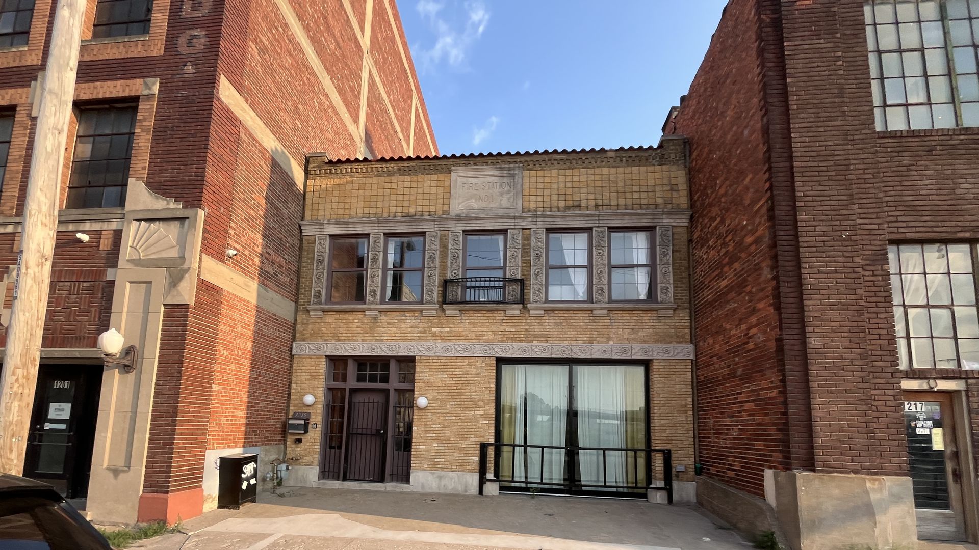 Two-story yellow brick building labeled Fire Station No. 1, flanked by taller red brick buildings, with windows, a black door, and a glass sliding door on the front under a clear blue sky.